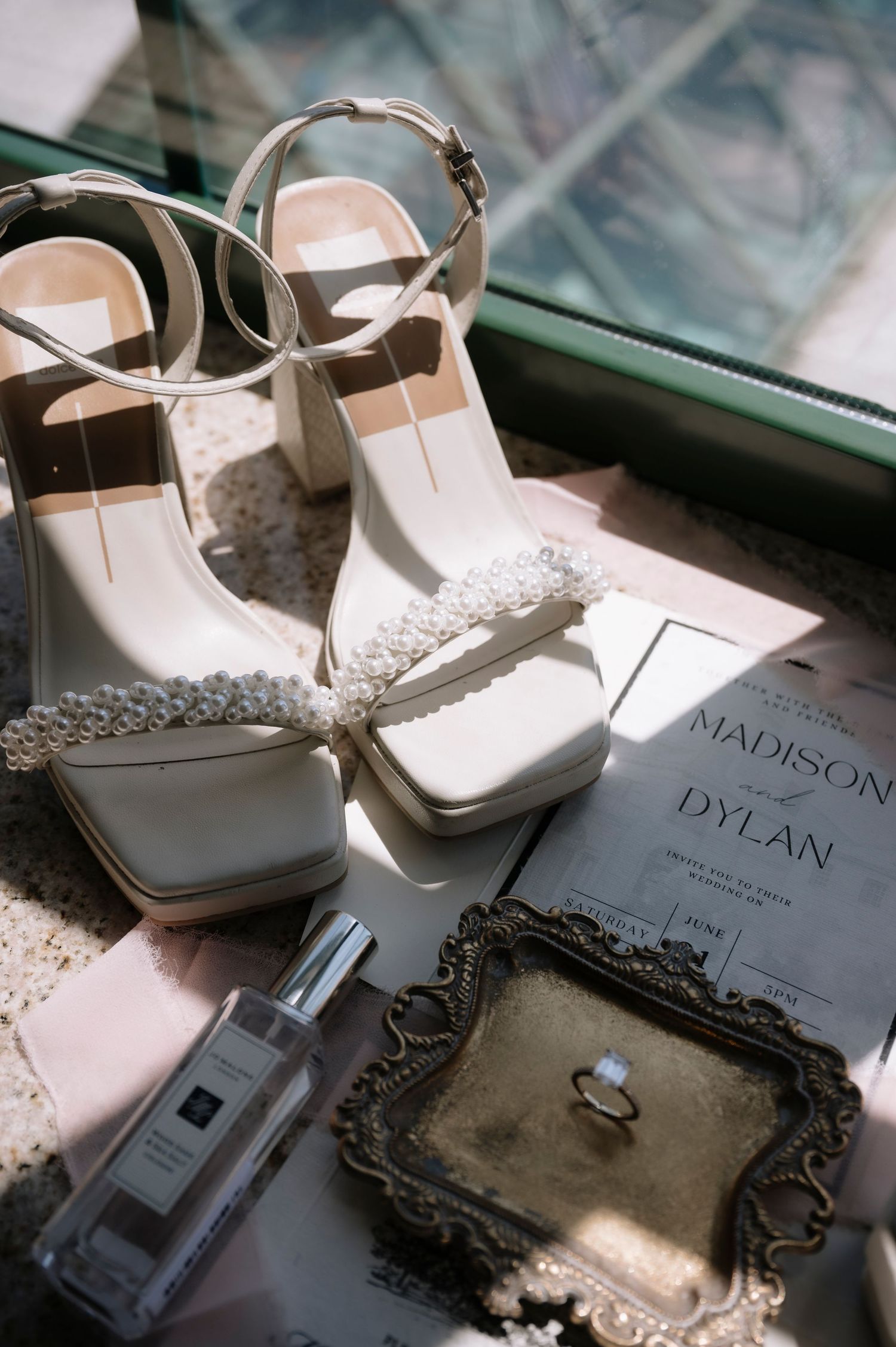 A wedding setup featuring white bridal shoes, perfume, an invitation, and an engagement ring in a decorative box on a reflective surface.