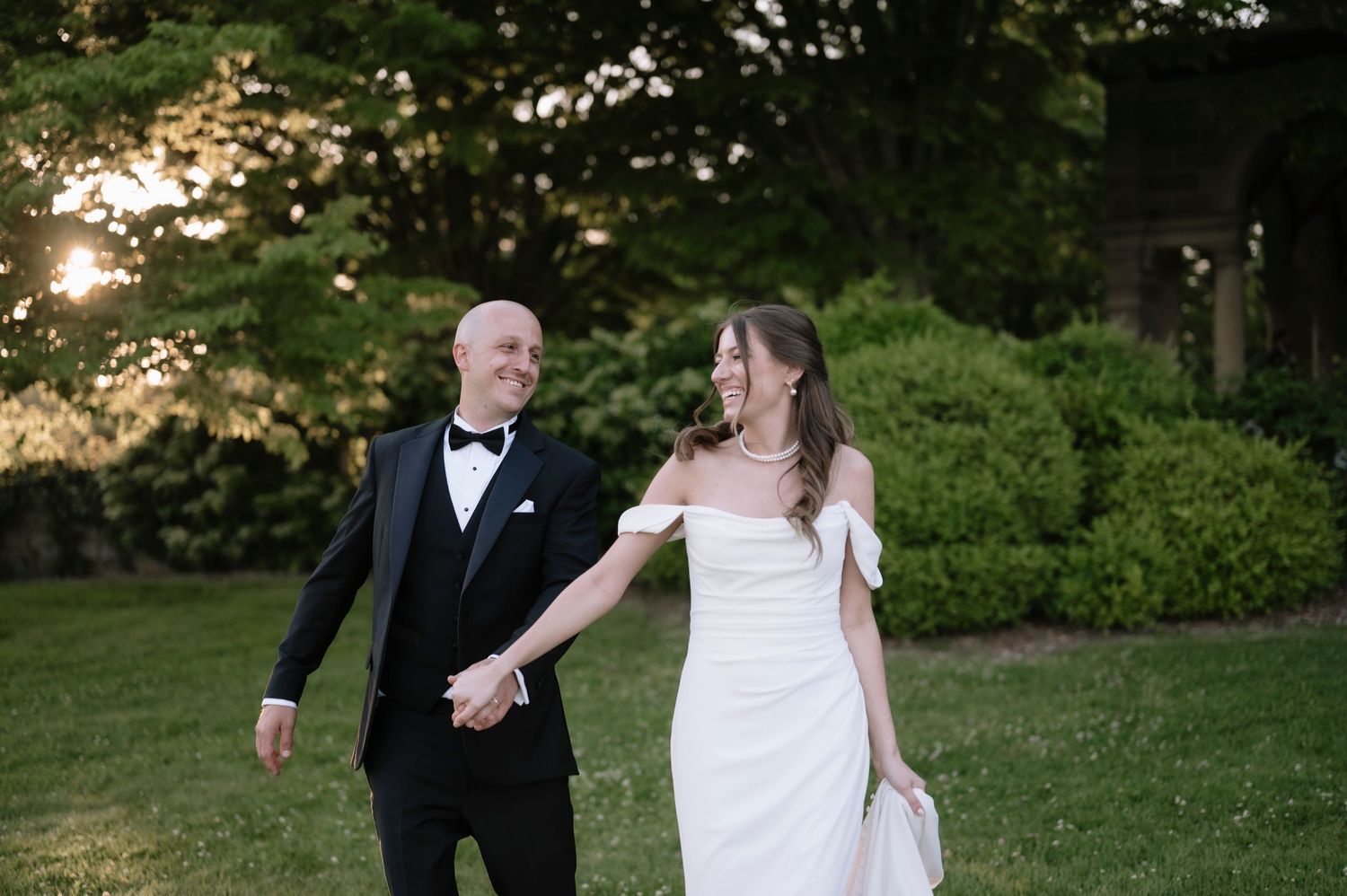Two people in formal wedding attire walk hand-in-hand across a lush green lawn at sunset.