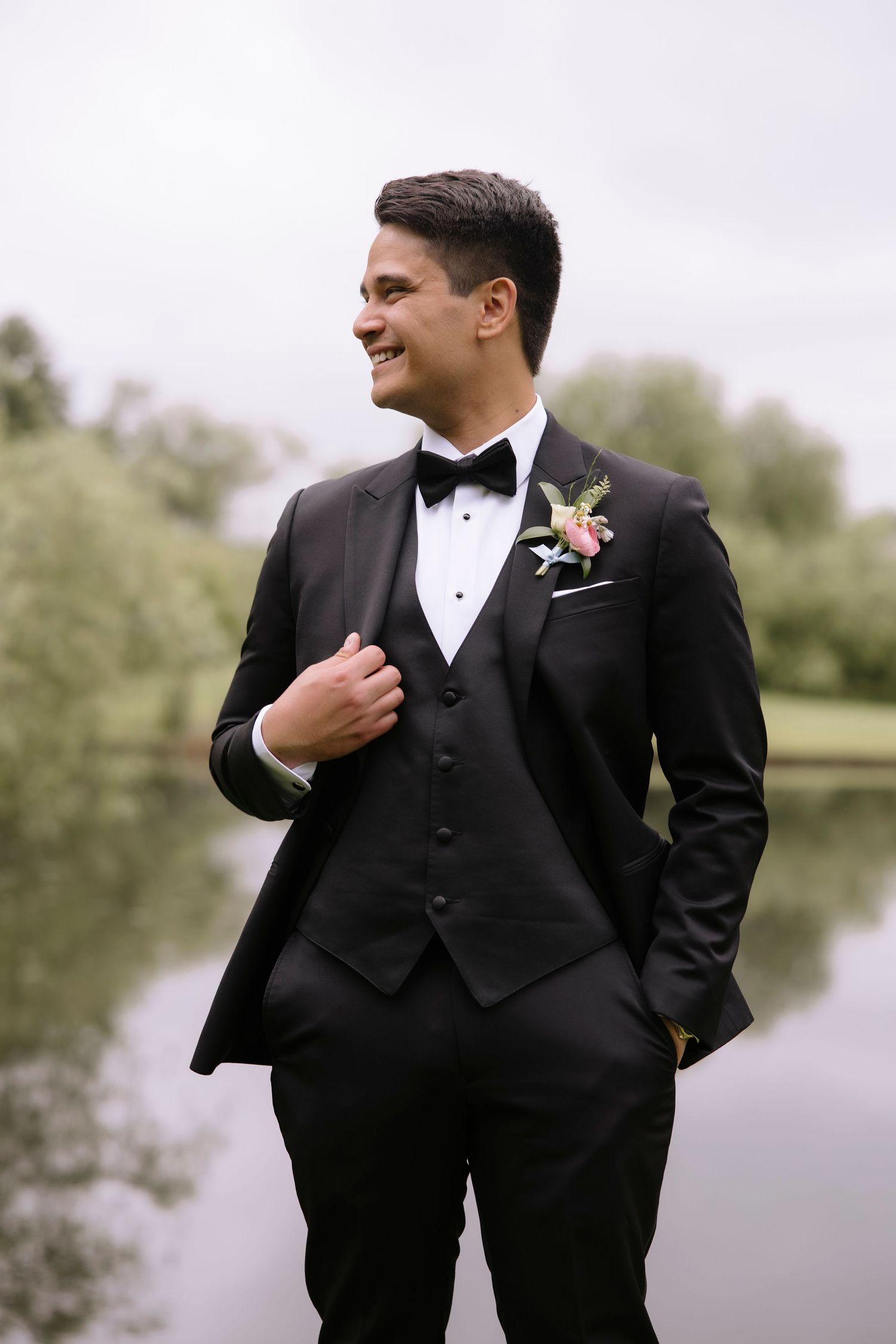 A groom in a classic black tuxedo with vest and bowtie stands smiling by a lake on his wedding day.