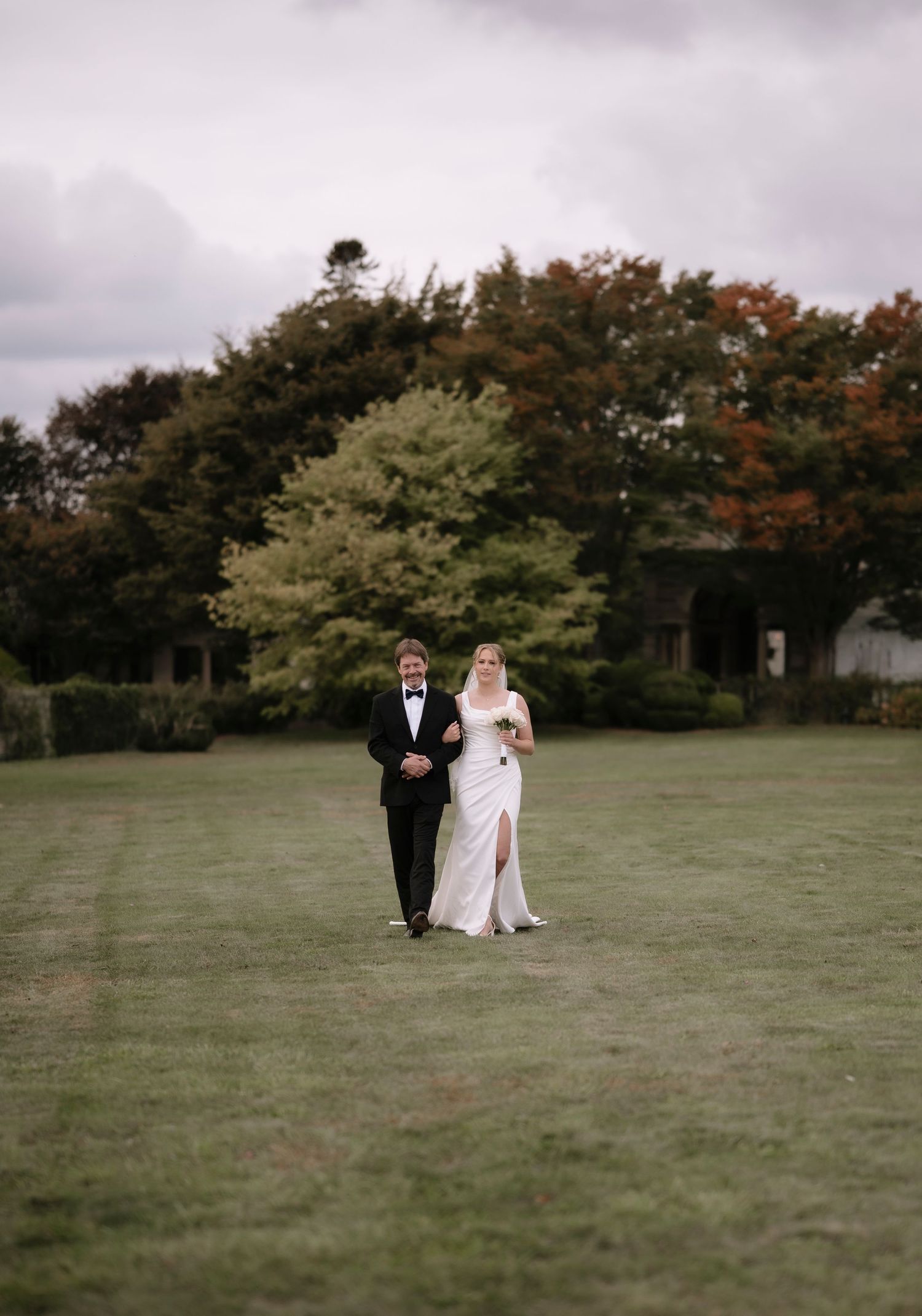 A couple in wedding attire walks together on a lush green lawn with autumn trees in the background.