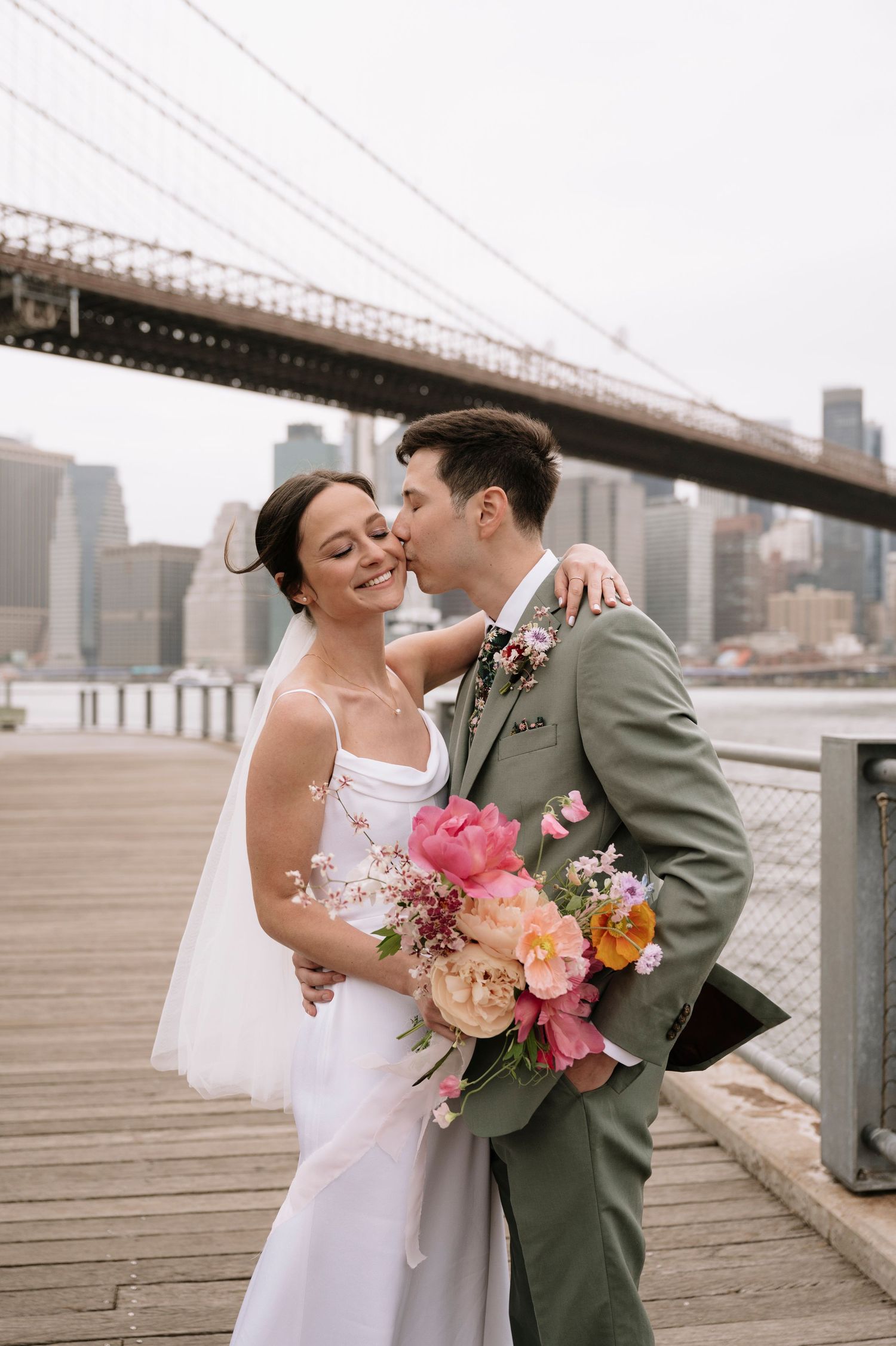 Newlyweds embracing with colorful bouquet by Manhattan Bridge on a cloudy day.