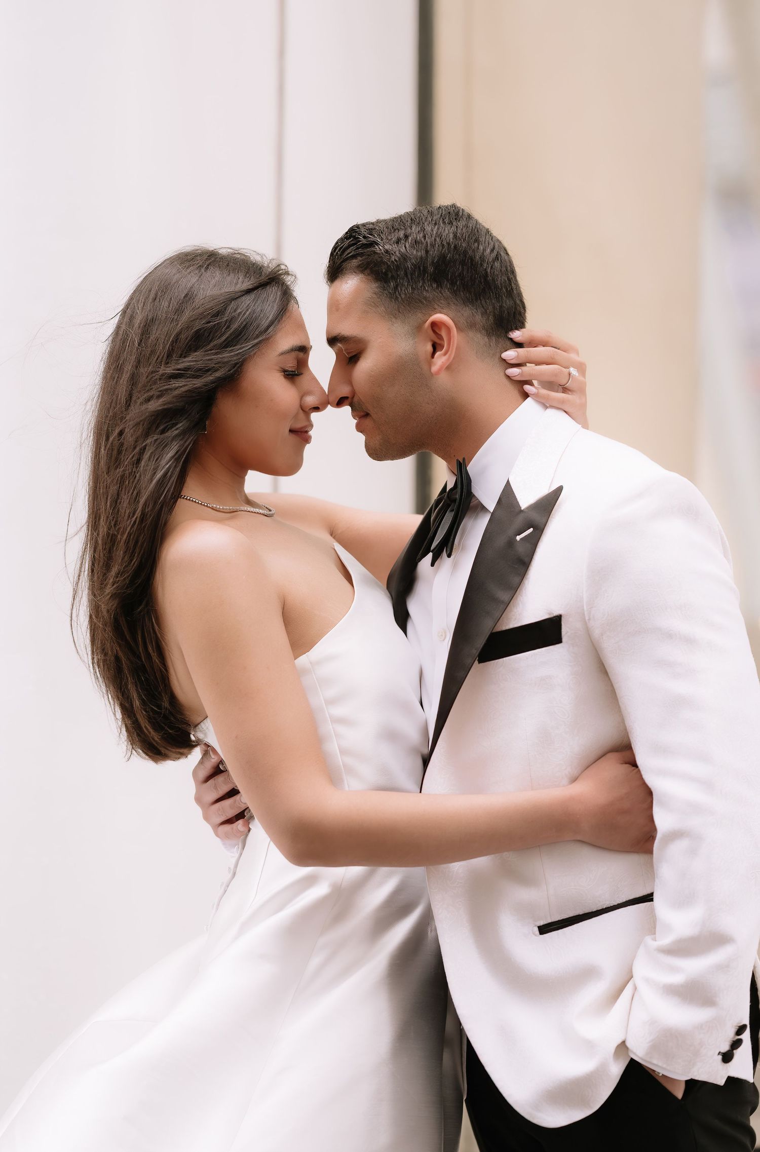 Romantic wedding couple in formal attire share an intimate moment against a light colored wall.