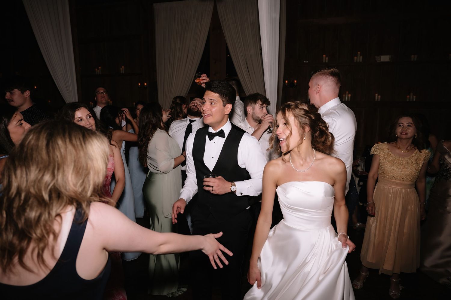 Bride in white strapless gown and groom in tuxedo dancing joyfully with wedding guests at reception.