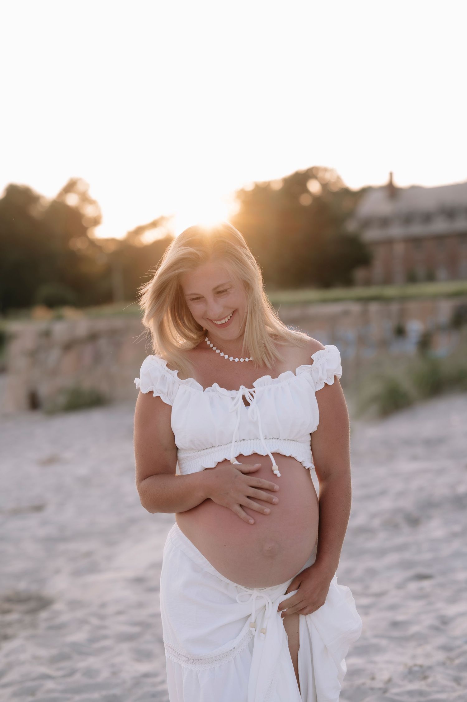 Pregnant woman in white crop top and skirt cradling baby bump on beach at golden sunset.