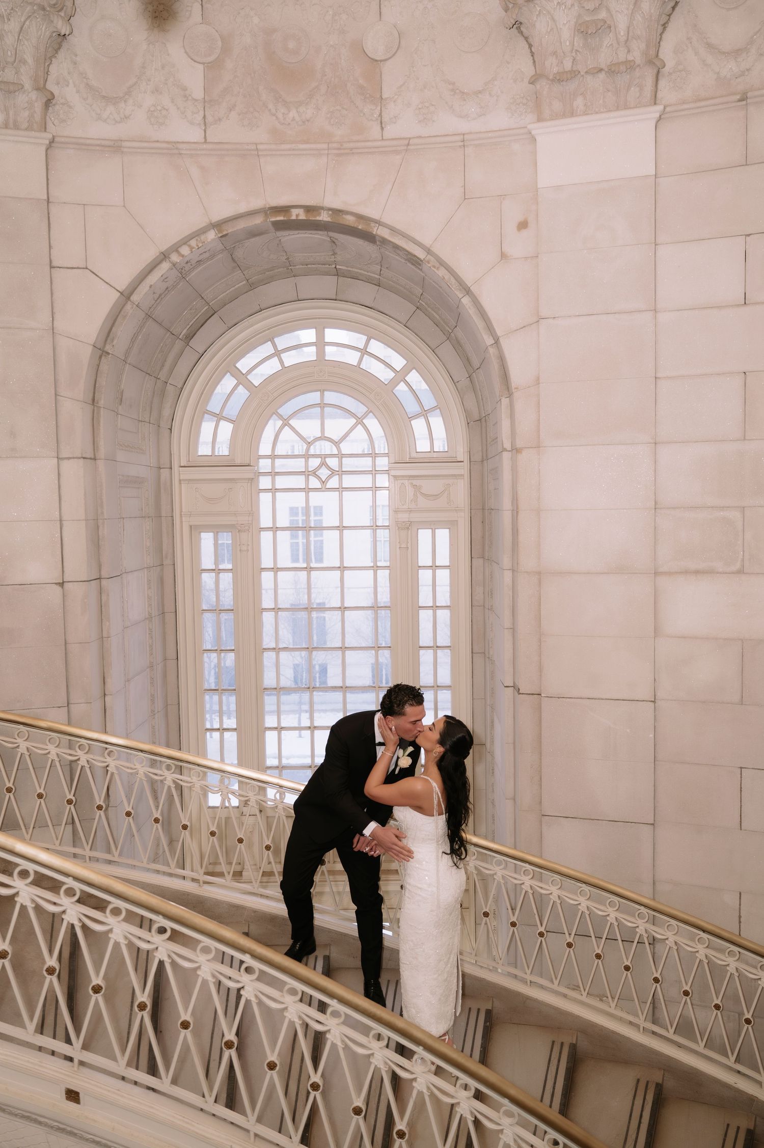 Bride and groom share a romantic kiss on an elegant staircase beneath an arched window in a grand marble venue.