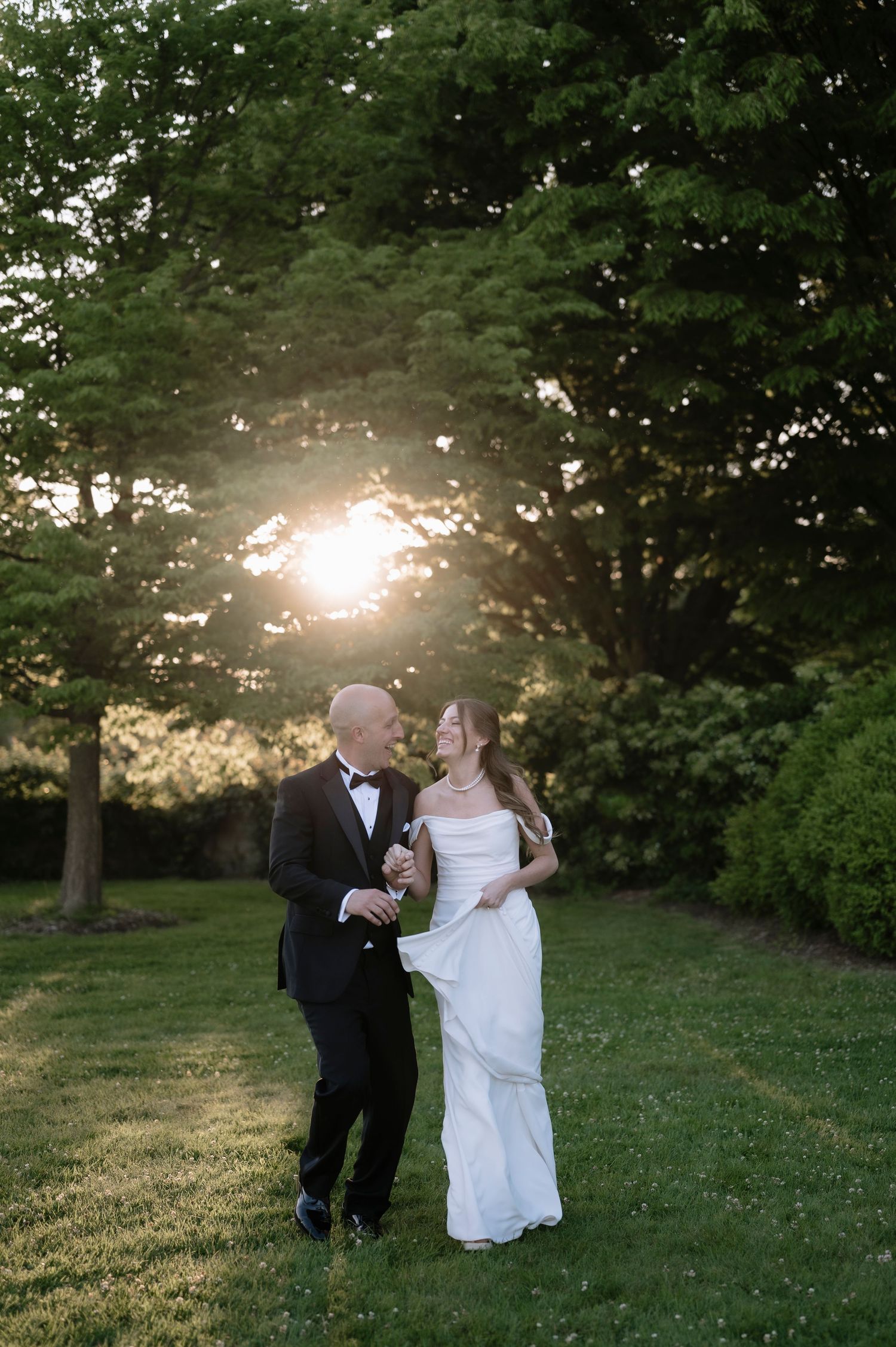 A couple in wedding attire walks together on a grassy lawn at sunset with trees creating a romantic backdrop.