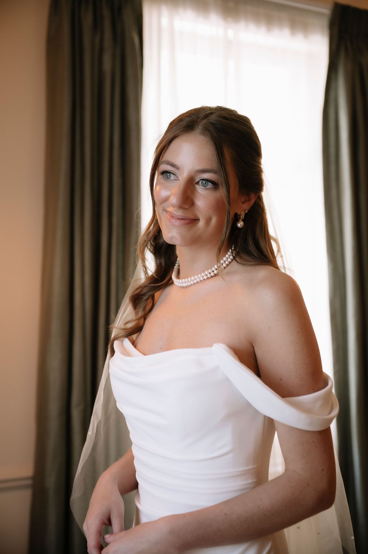 A bride wearing a classic white off-shoulder wedding dress and pearl necklace poses elegantly in front of curtains.