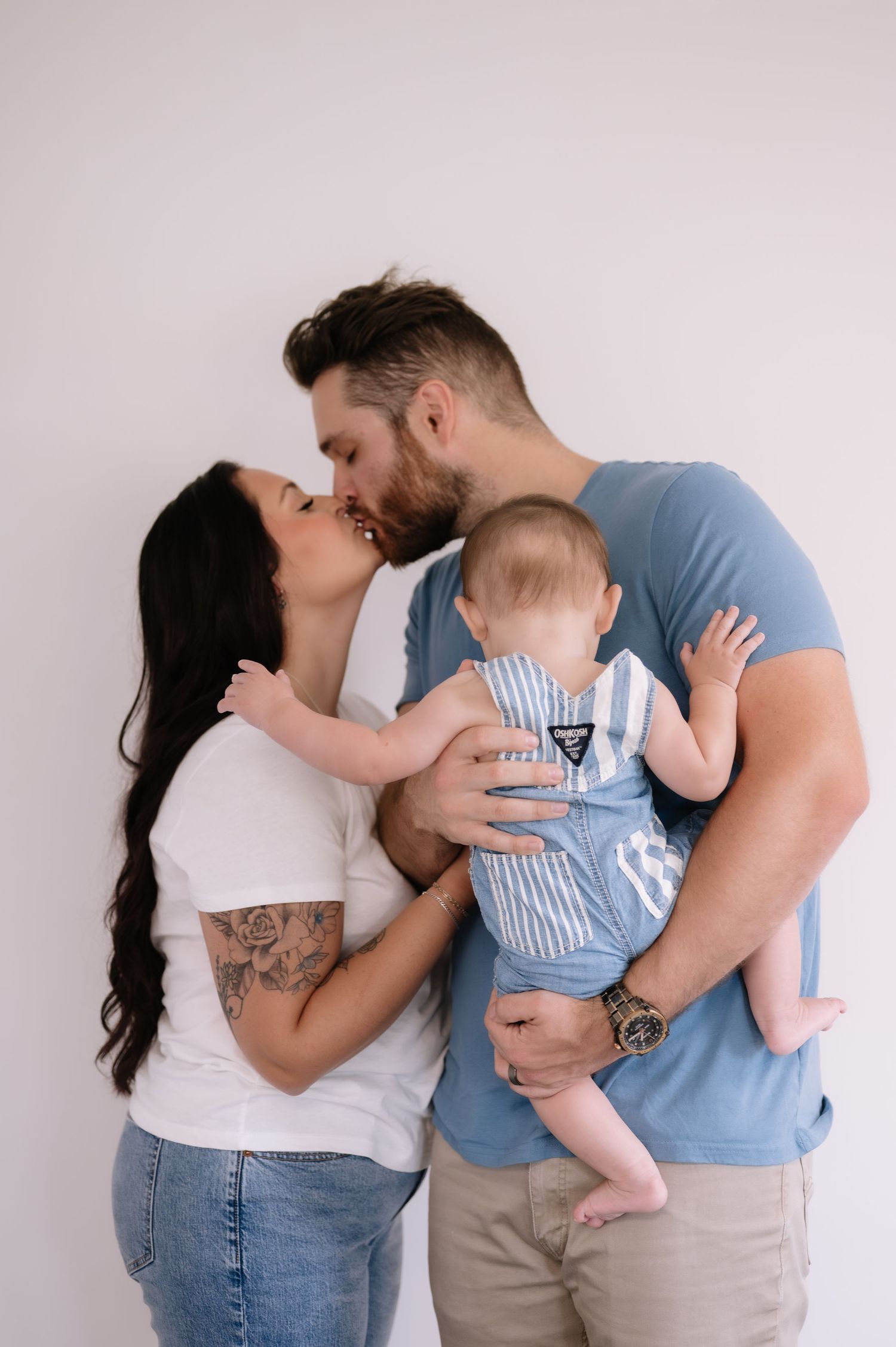 A loving couple shares a tender kiss while holding their baby in striped overalls against a light background.