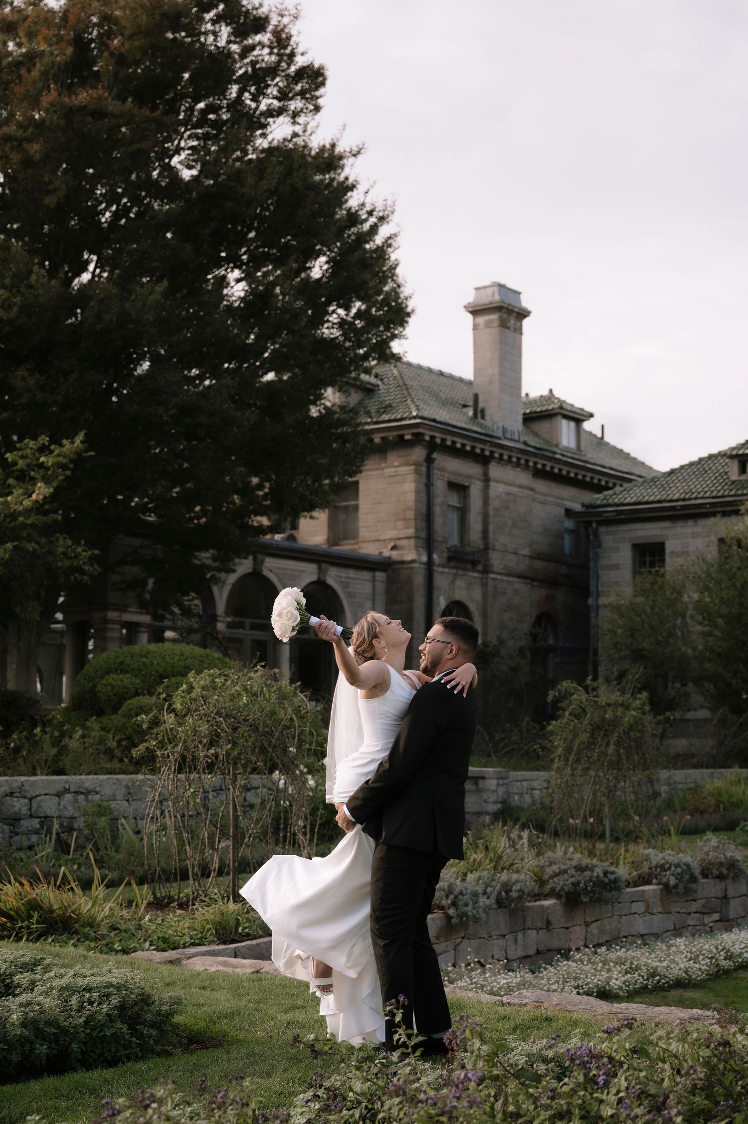 Bride and groom dance together in a garden setting with historic stone building in background.