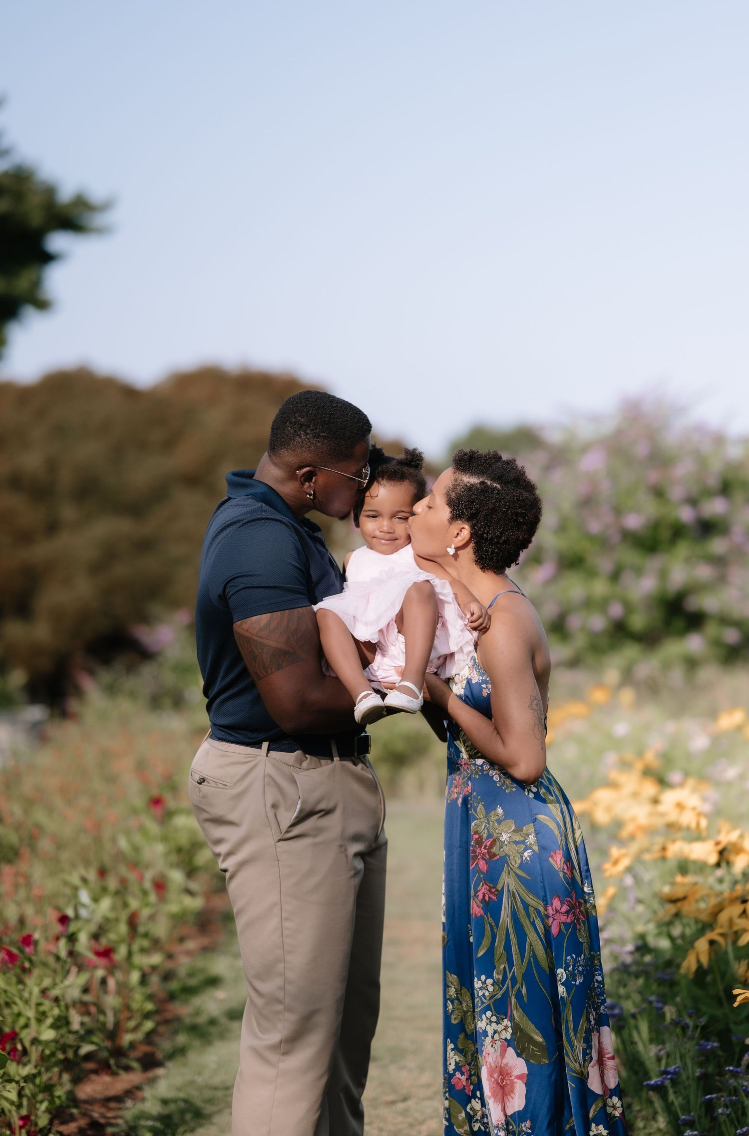 A loving family of three poses in a colorful flower garden, with parents kissing their baby girl on the cheeks.