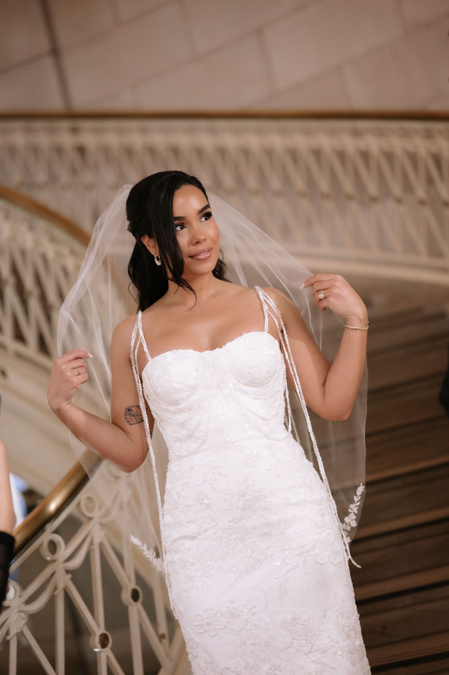 Bride in fitted lace gown and veil poses gracefully on elegant staircase, smiling softly in a classic wedding portrait.