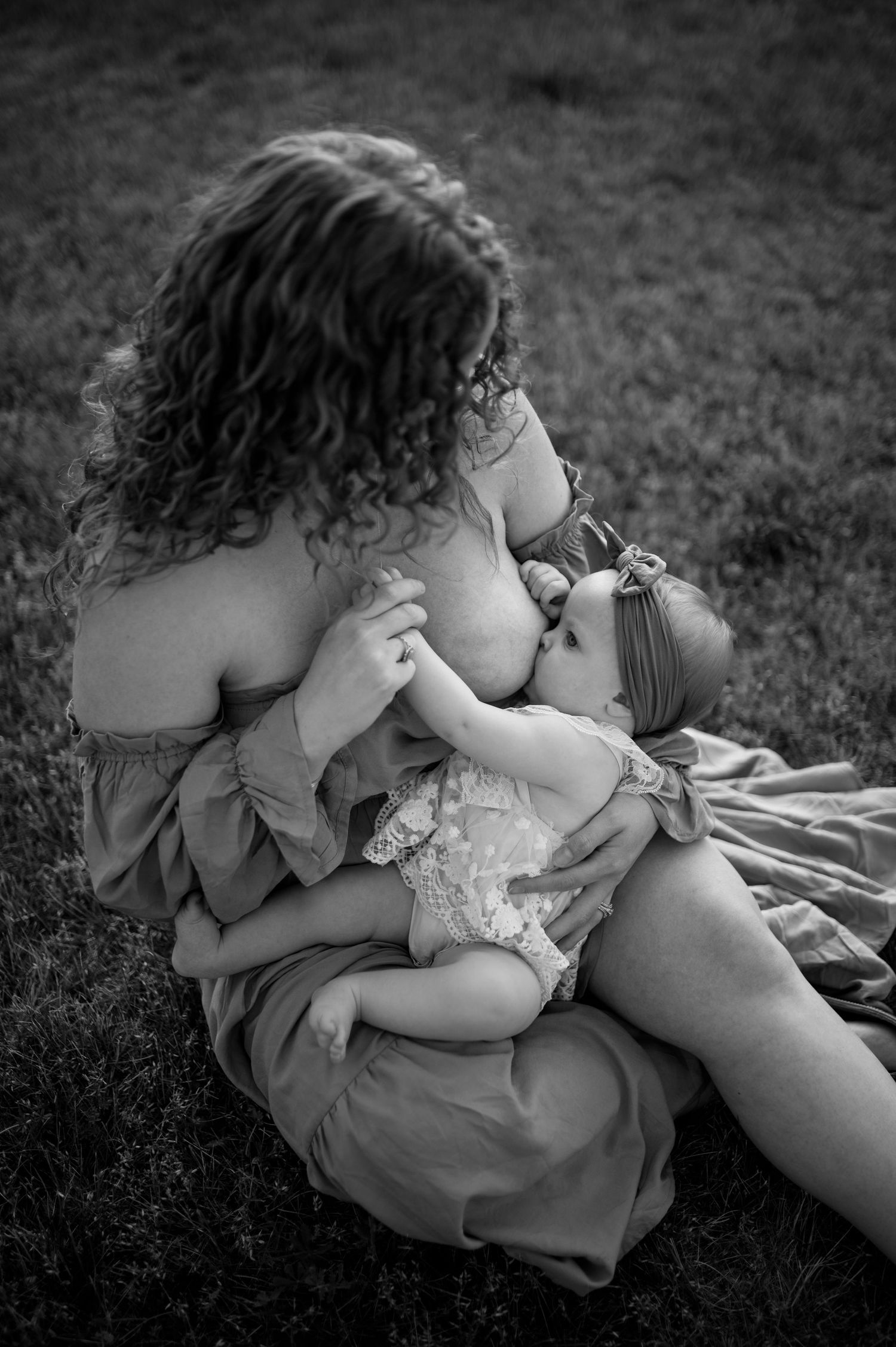 Intimate black and white photograph of mother nursing baby outdoors on grass in flowing dress.