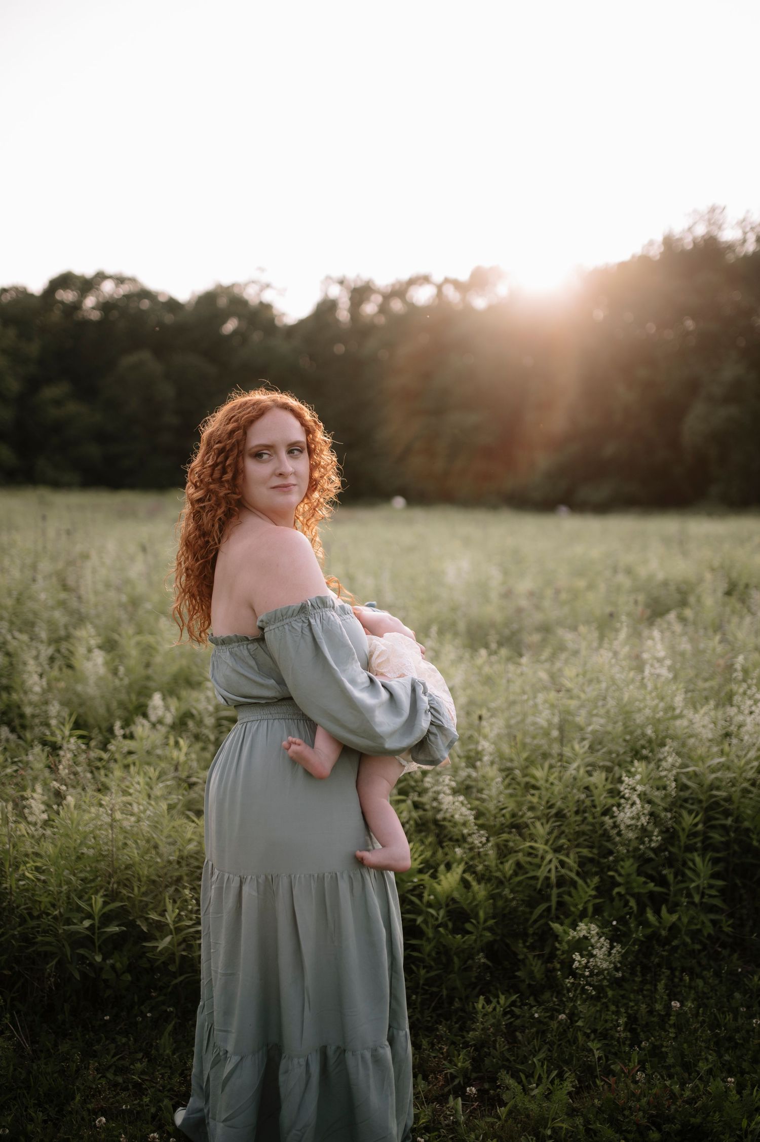 Person in flowing sage green dress stands in sunlit meadow during golden hour with sunflare in background.