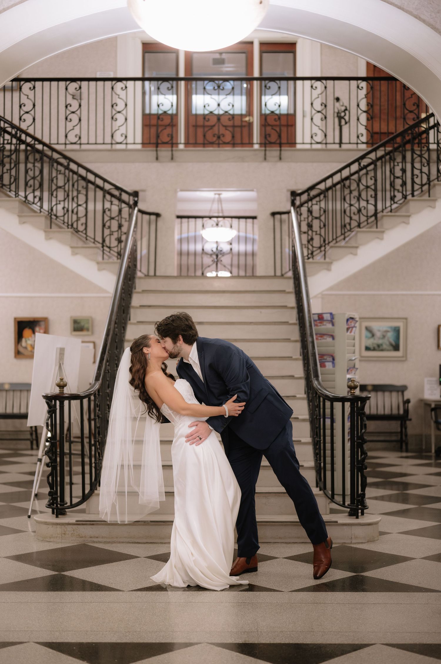 Couple shares romantic dance pose on checkerboard floor beneath arched staircase.