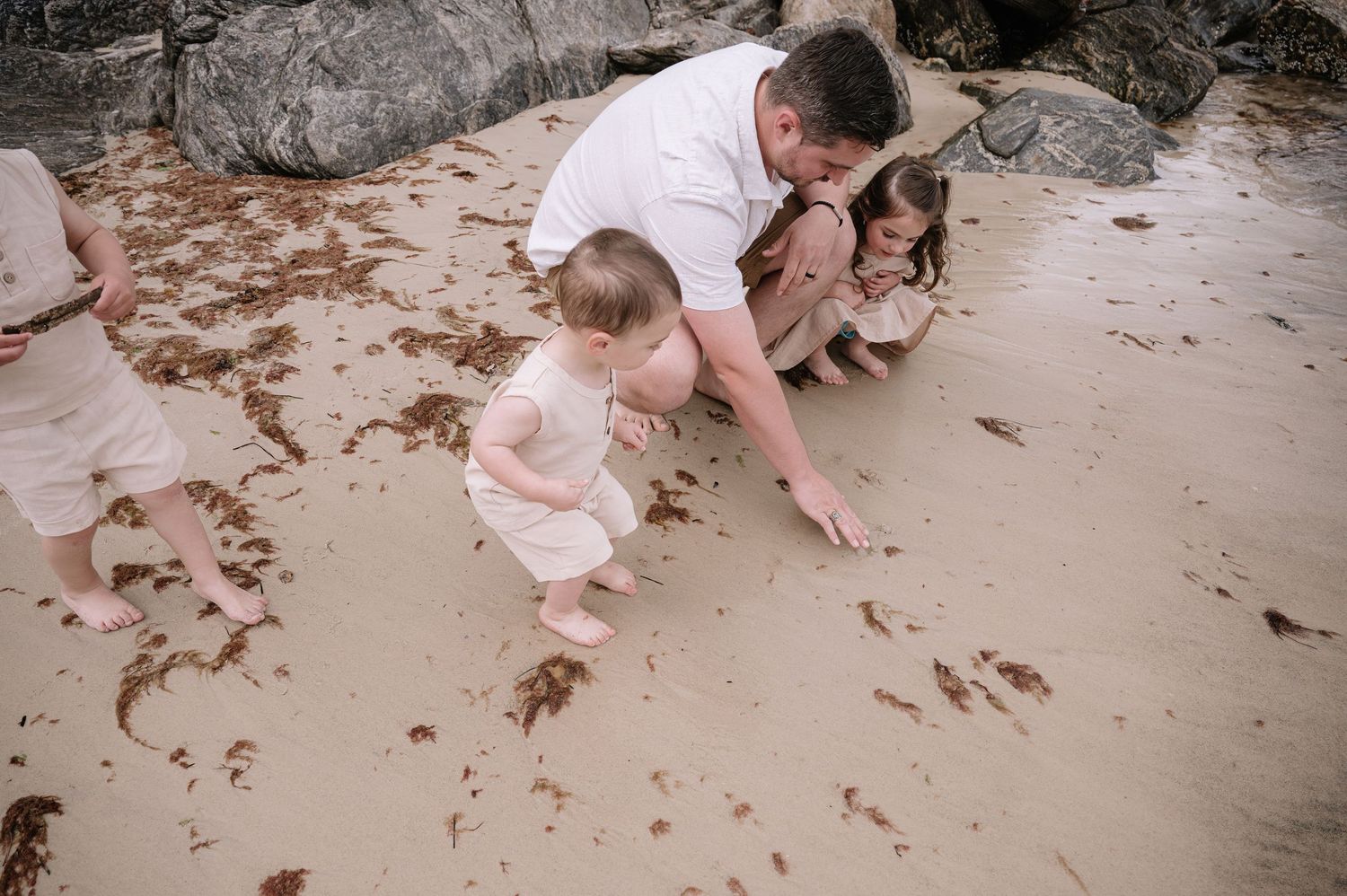 People and a brown dog sit together on a sandy beach near rocky outcrops.