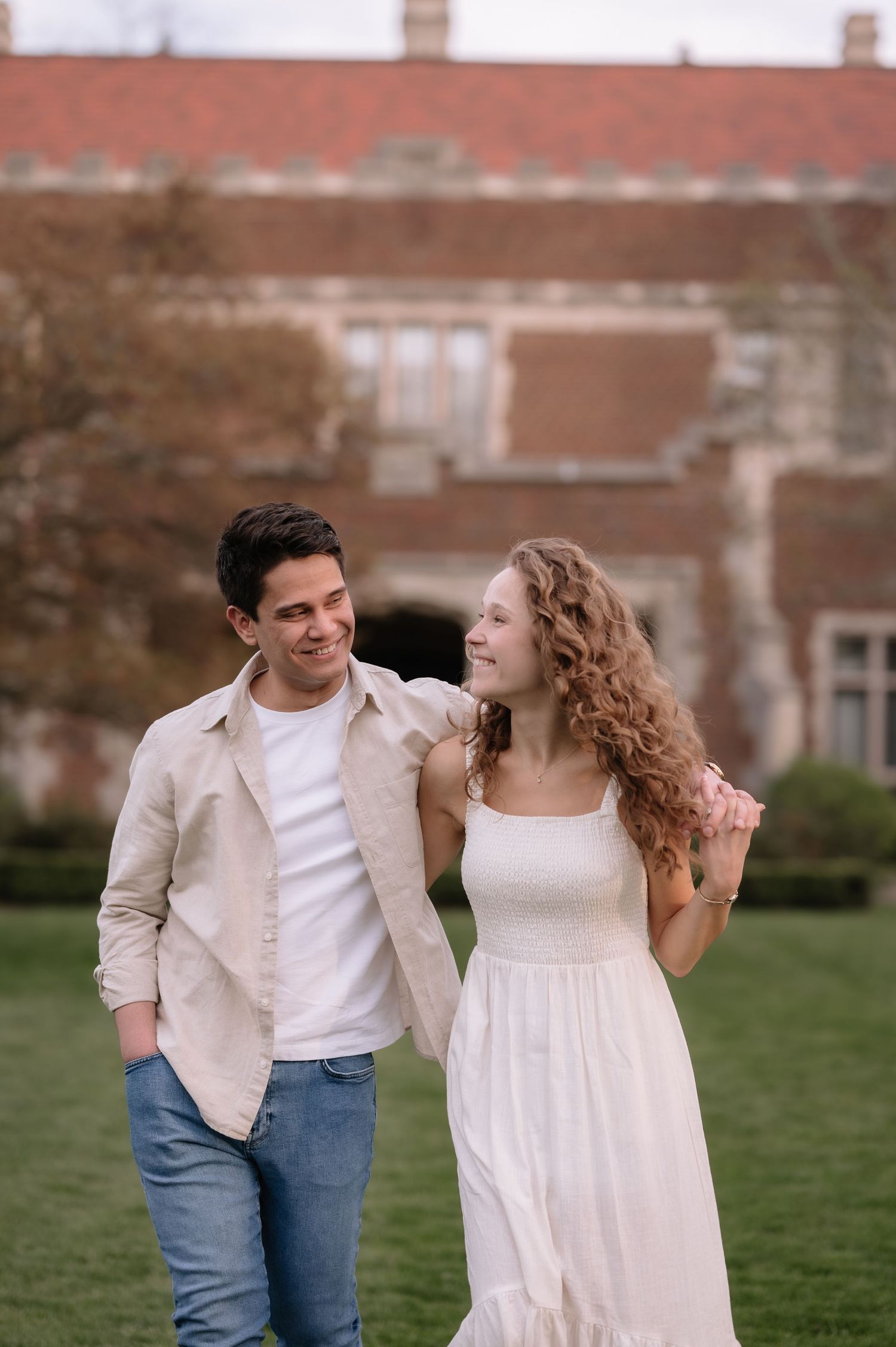 A casual couple shares a lighthearted moment on a green lawn with historic buildings in the background.