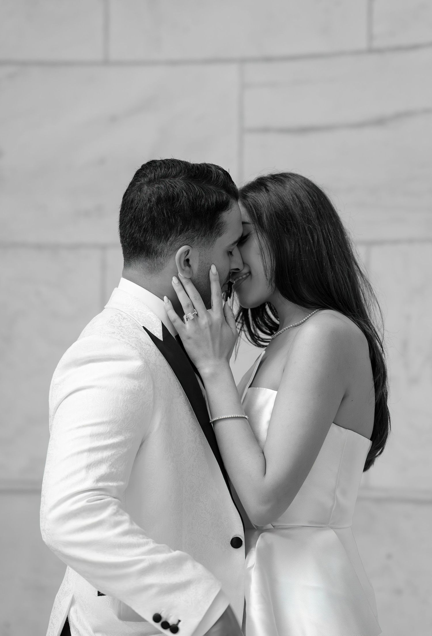 A romantic black and white photograph of a couple in elegant white outfits sharing an intimate moment against a light wall.