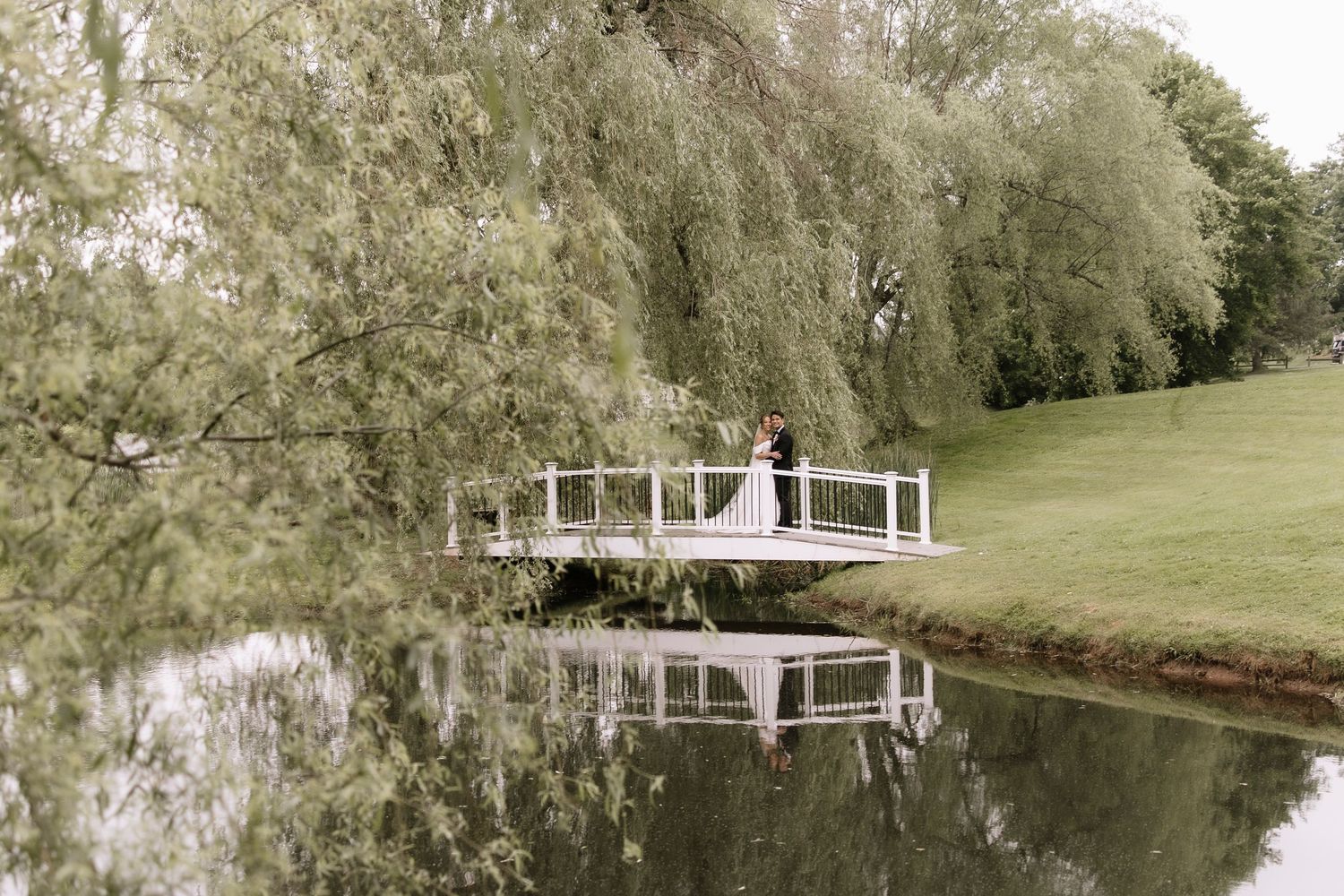 Bride and groom share a romantic kiss on a white bridge over a calm pond surrounded by weeping willows.
