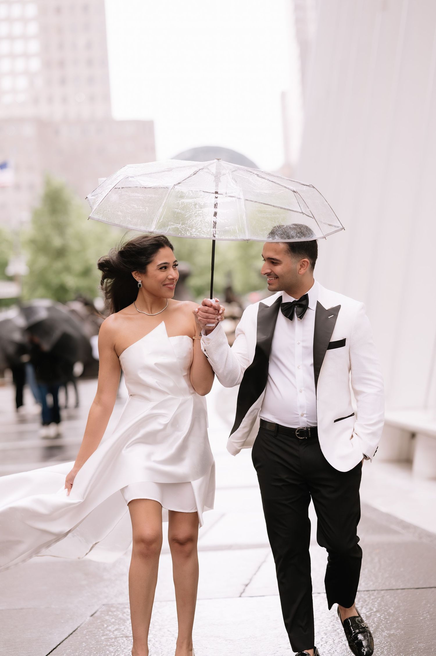 Joyful newlyweds walking in the rain sharing a clear umbrella in an urban setting.