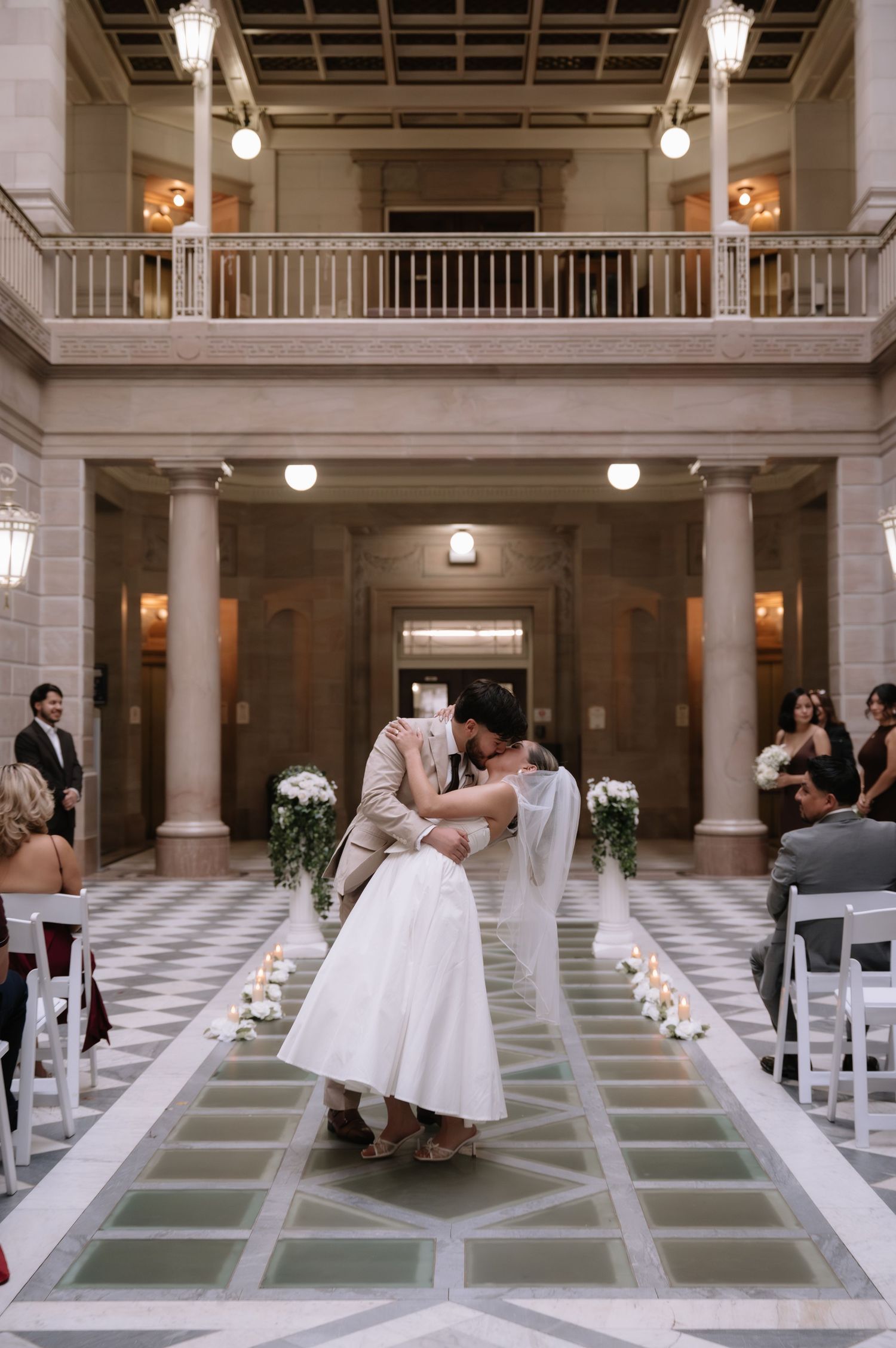 A wedding ceremony setup in a grand marble atrium with white chairs arranged in rows and elegant lighting fixtures above.