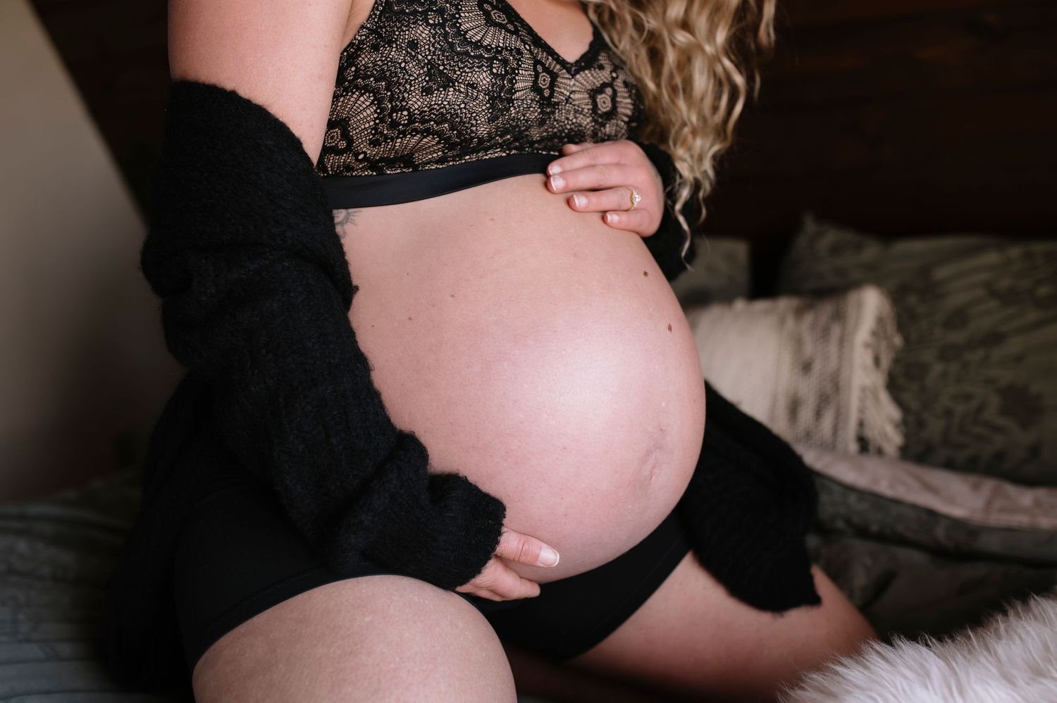 A pregnant woman in black lace lingerie poses for a maternity photoshoot with dramatic lighting.