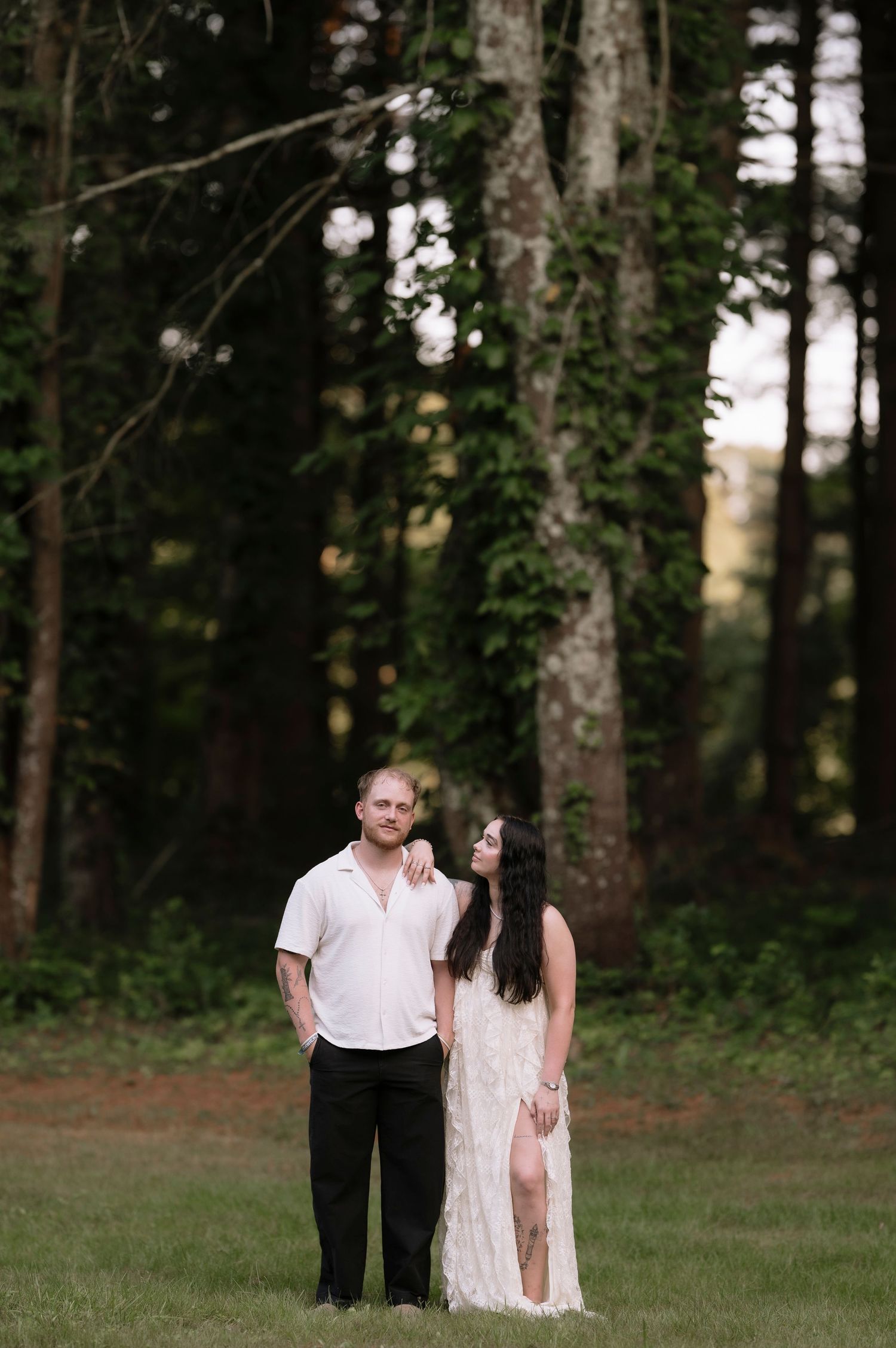 Couple standing together in a lush green forest setting, woman in white lace dress beside man in white shirt.