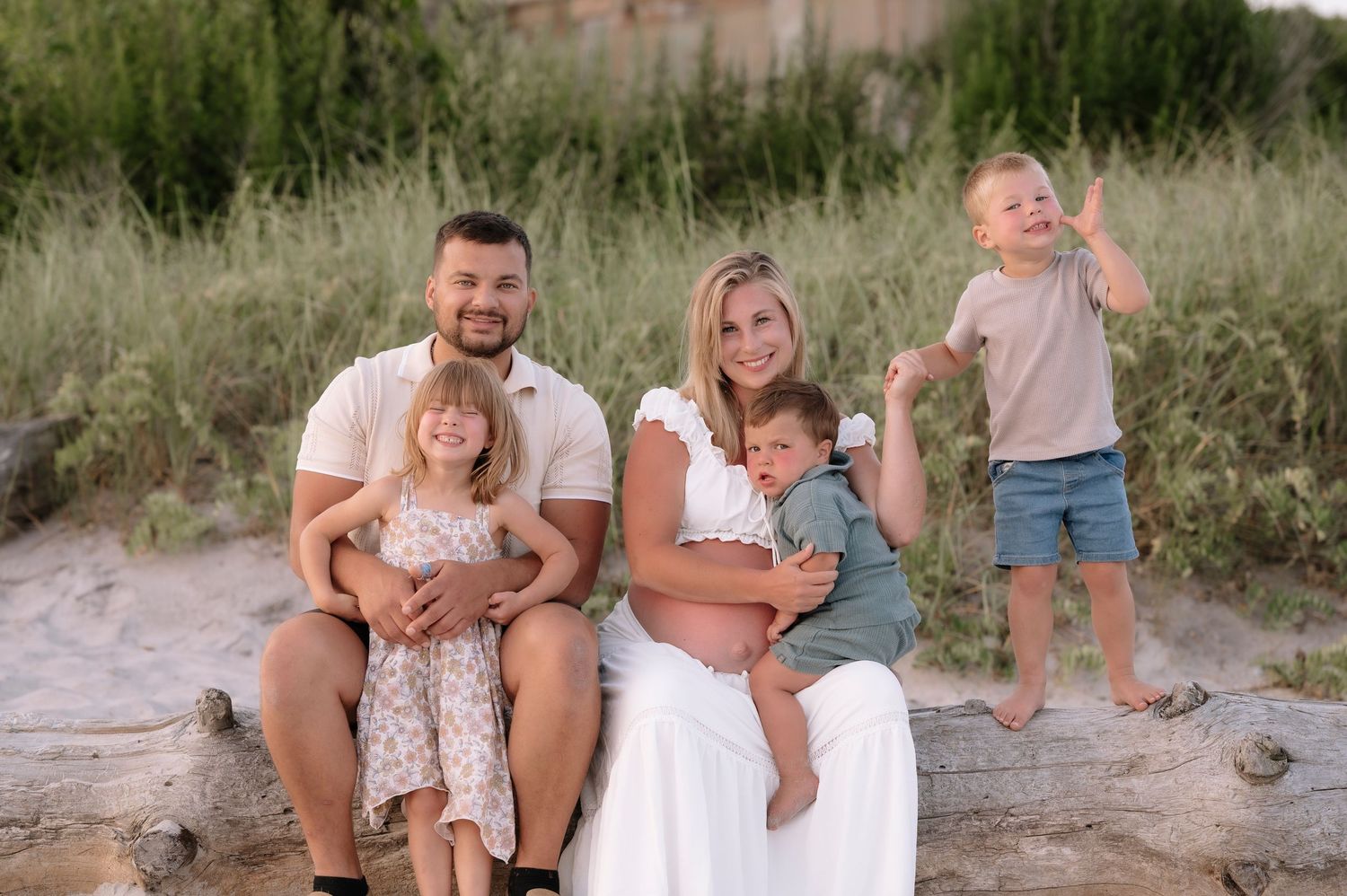 Happy family of five posing outdoors on sandy beach with lush green dunes, parents with three young children smiling.