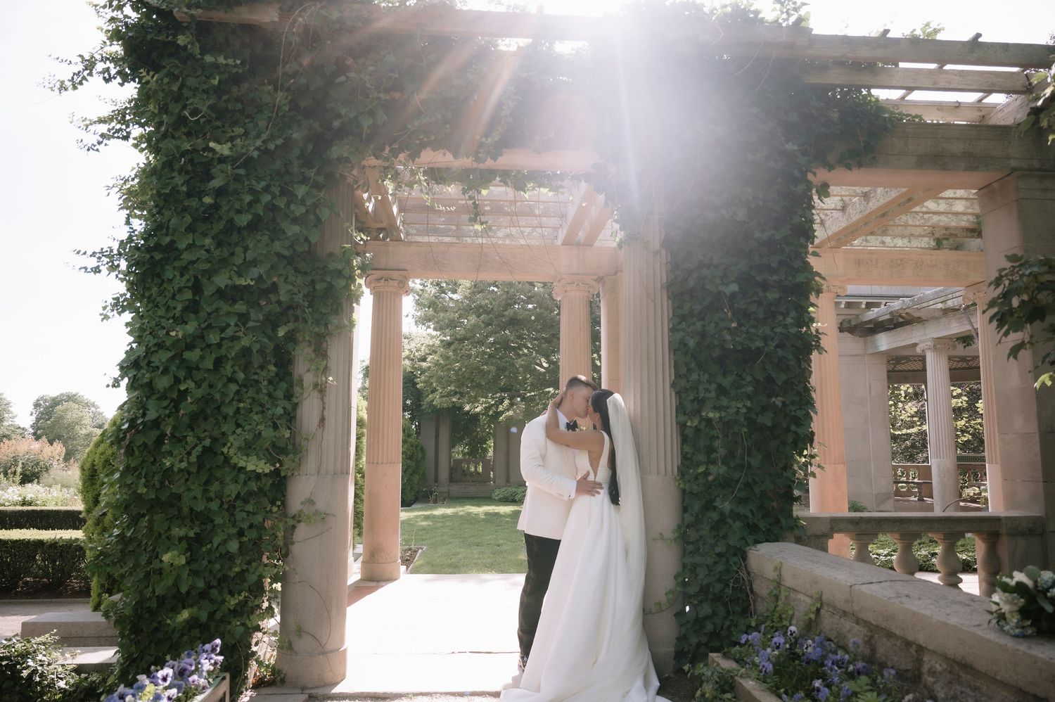 Bride and groom share romantic kiss under ivy-covered stone pergola arch in sunlit garden wedding ceremony.