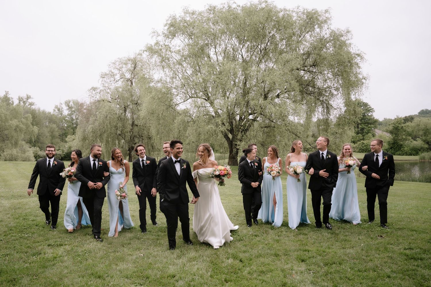 Wedding party in formal attire walks across green lawn with large willow tree in background.
