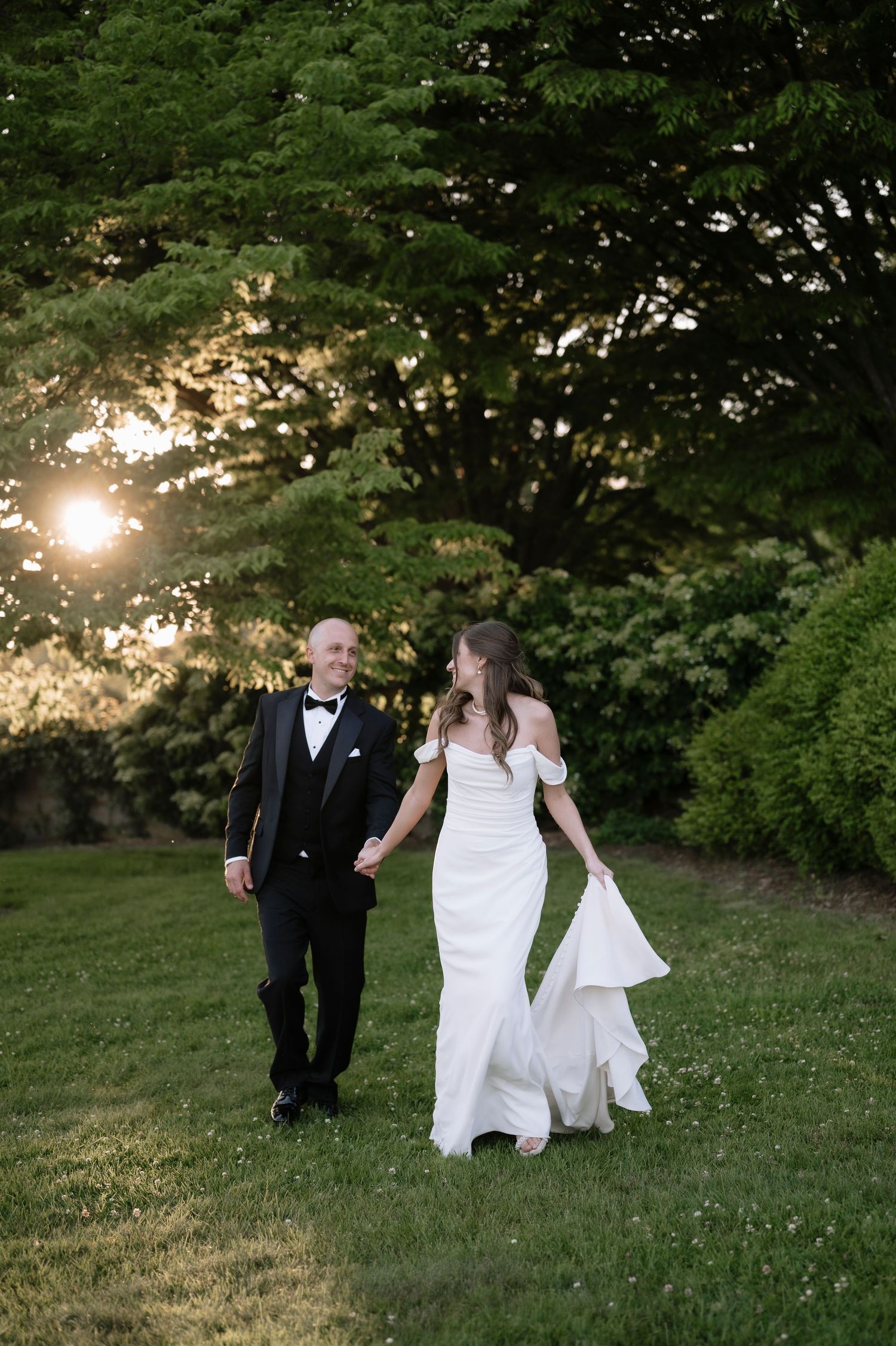 A couple in wedding attire walks hand in hand on a grassy hillside at sunset with lush green trees in the background.