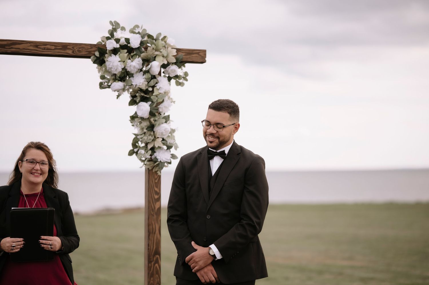 A couple exchanges vows during an outdoor wedding ceremony beneath a wooden cross adorned with white flowers.