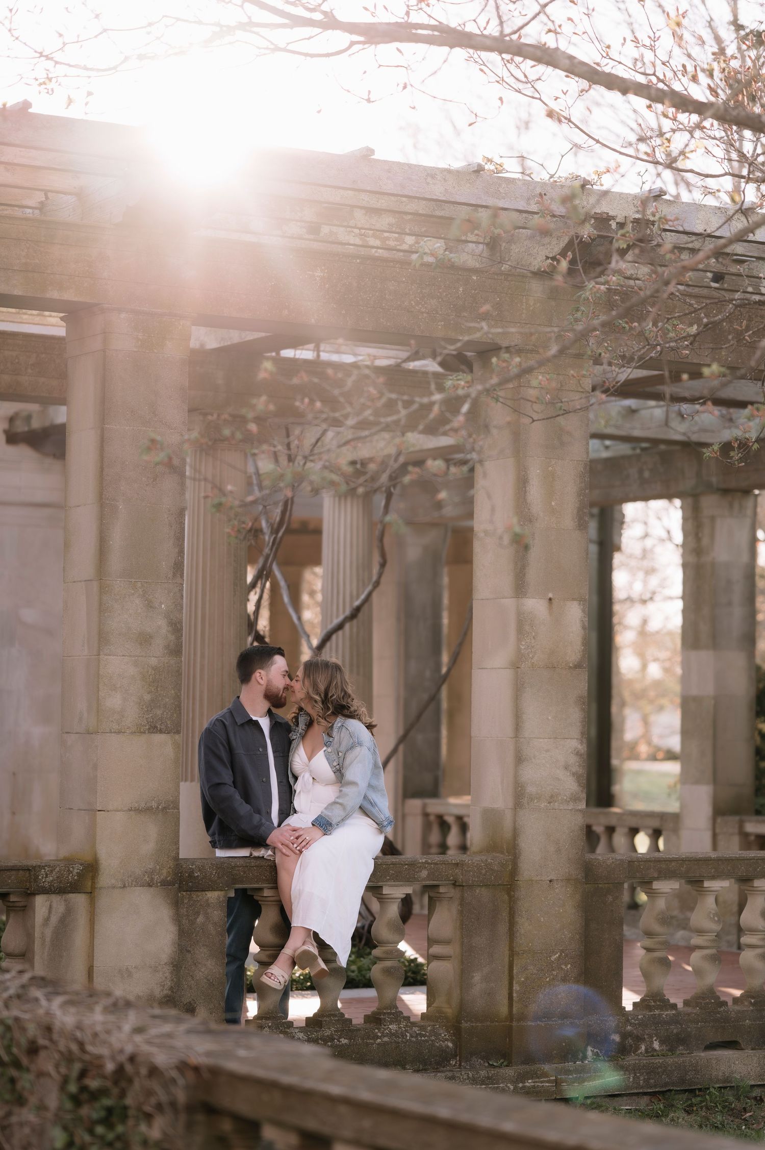 Couple sharing a romantic kiss on a stone balustrade at a sunlit pergola during an outdoor engagement session.