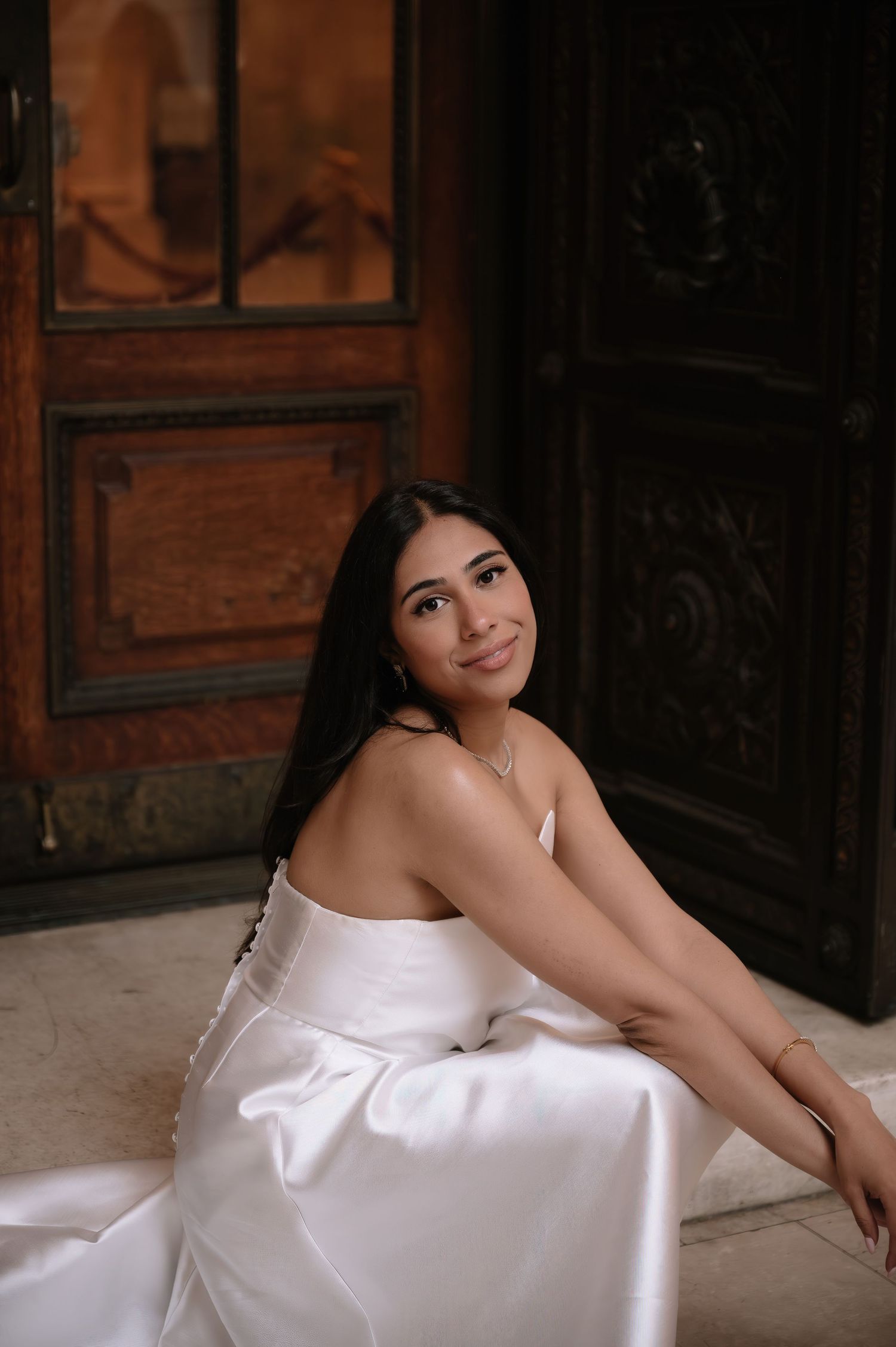 A person in a white strapless dress sits gracefully against ornate wooden doors in a romantic portrait setting.
