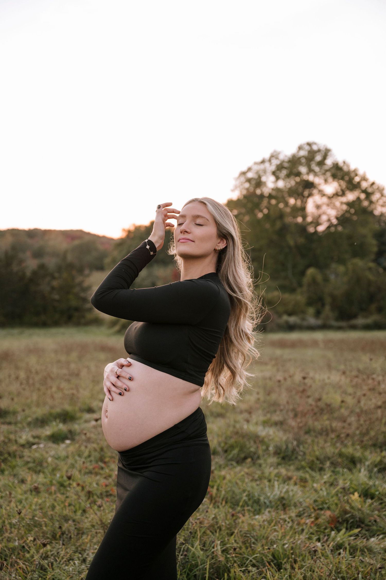 An expectant mother in black clothing poses gracefully in a sunlit autumn field.