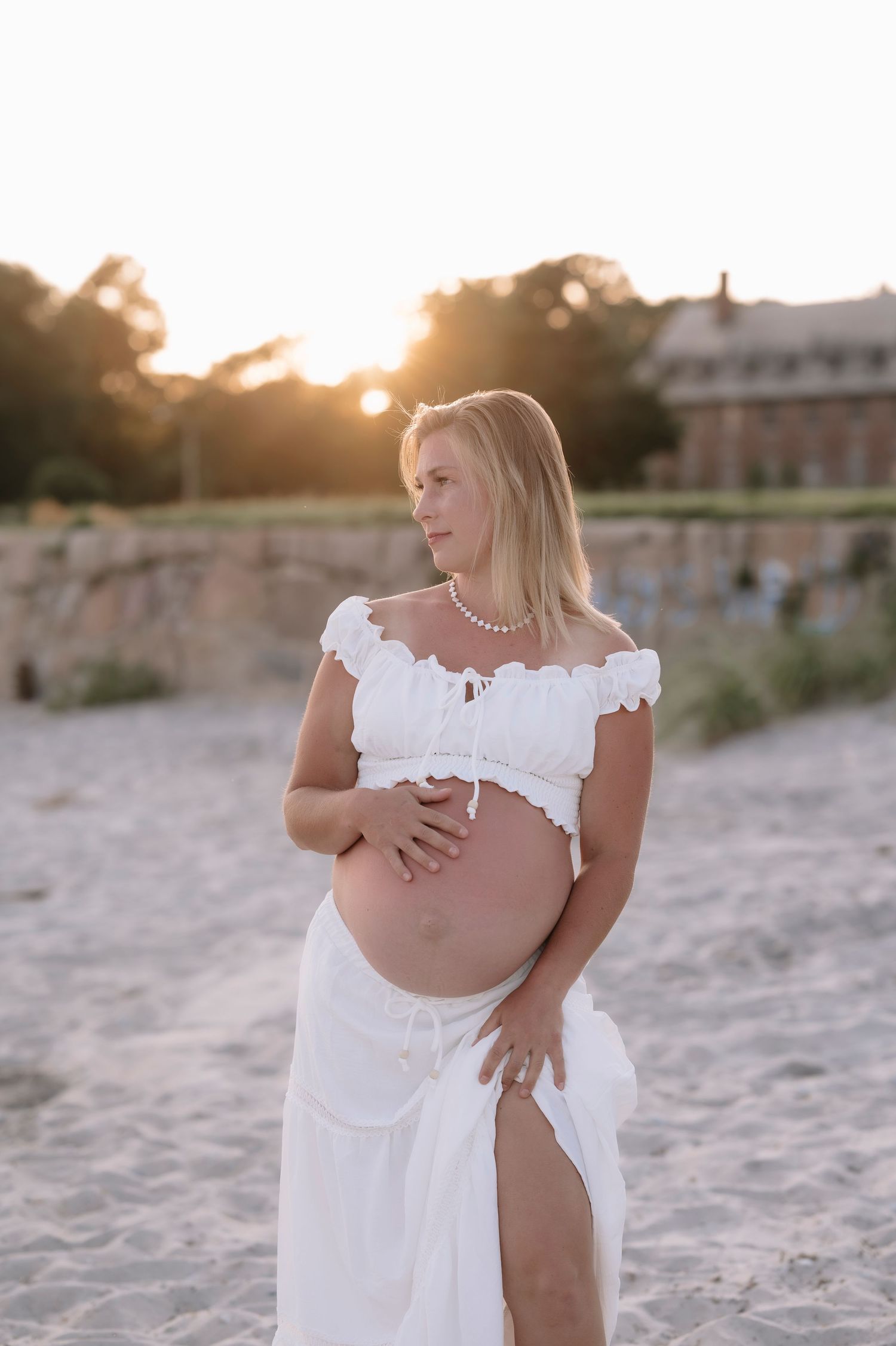 Blonde pregnant woman in white off-shoulder crop top and skirt cradles baby bump on sandy beach at golden hour.