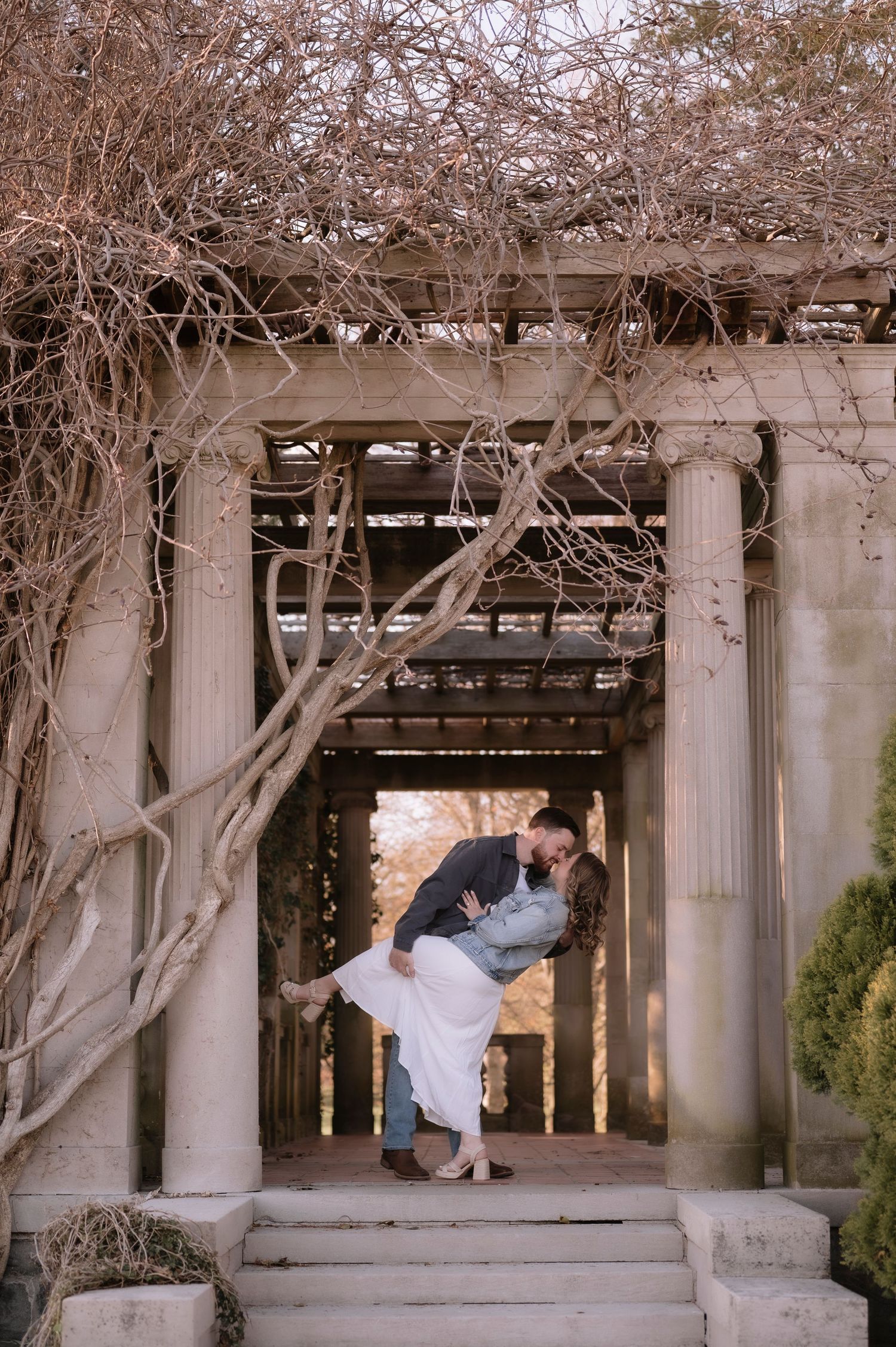 Couple sharing a romantic dip kiss under a stone pergola with bare winter vines during an engagement session.