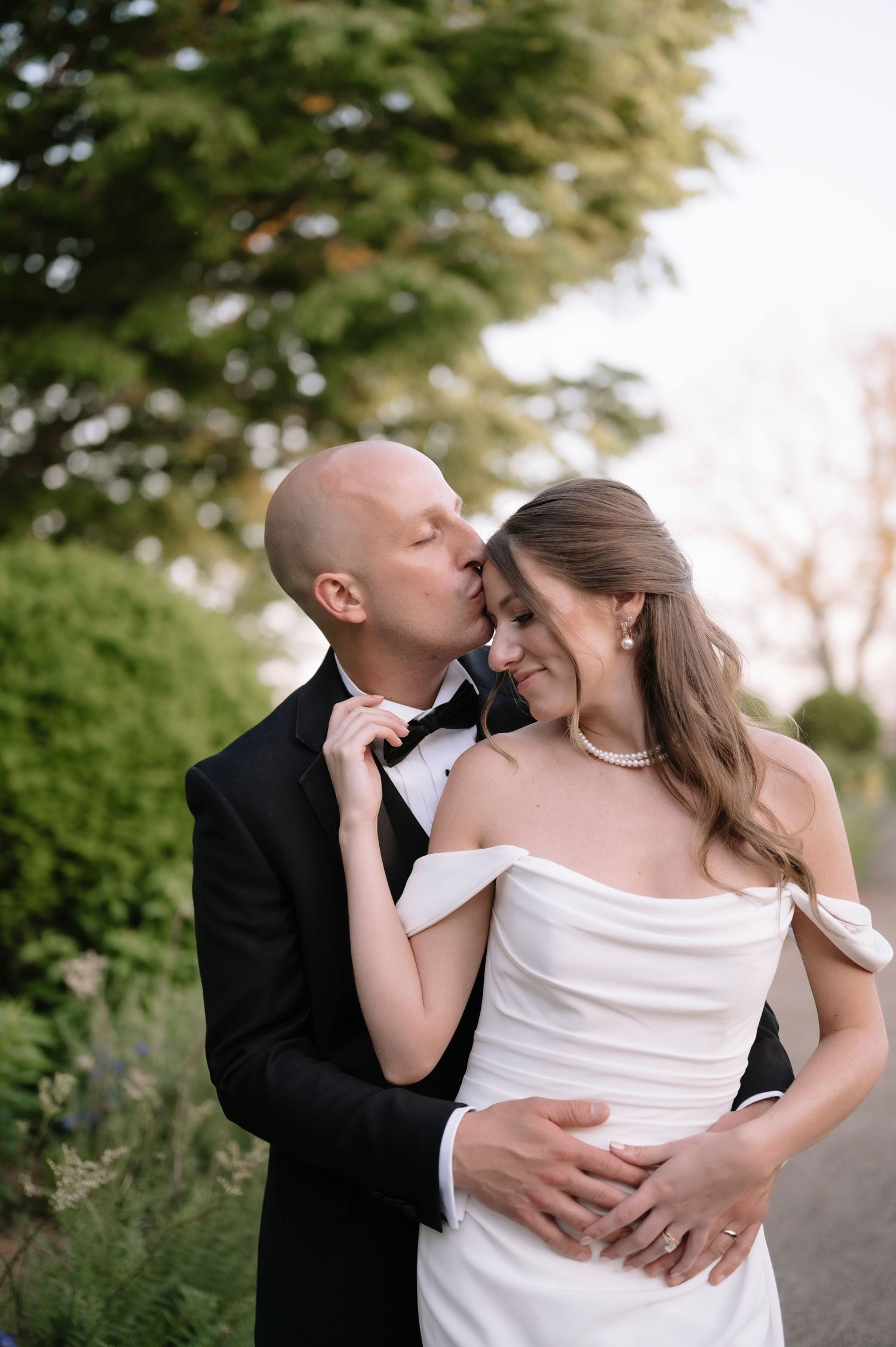 A romantic outdoor wedding photoshoot shows an intimate moment between newlyweds on a tree-lined pathway.