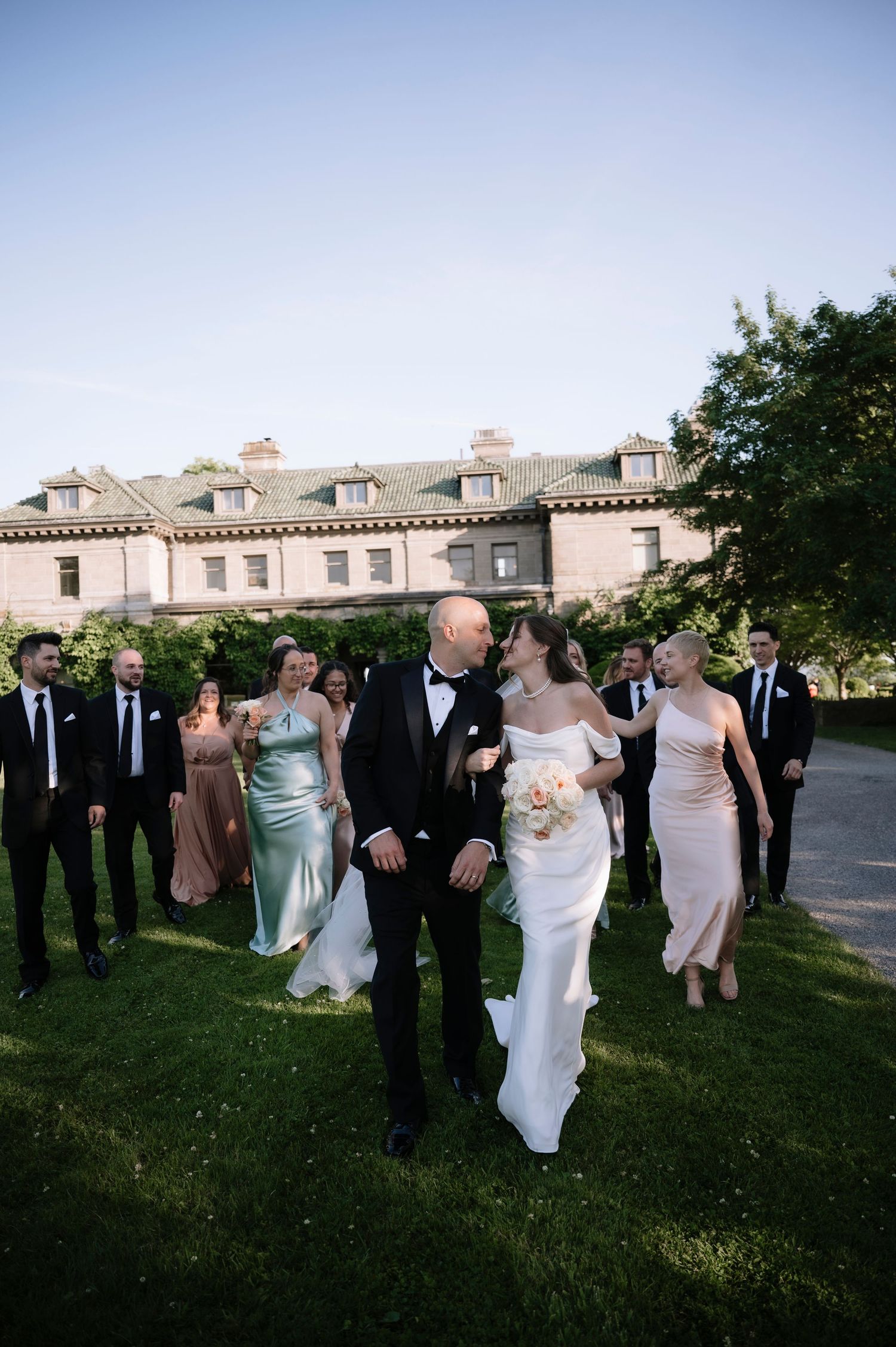 Wedding party walks together outdoors with formal suits and dresses in a casual outdoor setting at dusk.