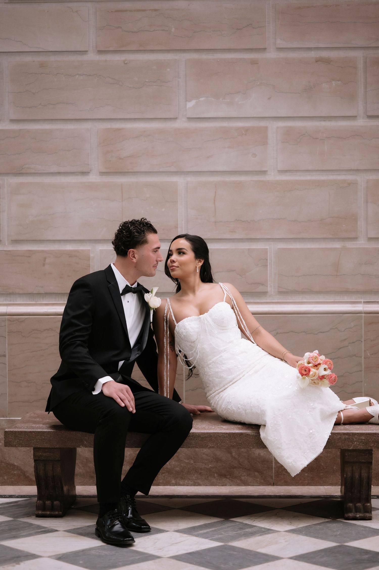 Bride and groom sit together on a marble bench, sharing a romantic gaze, dressed in a white gown and black tuxedo.