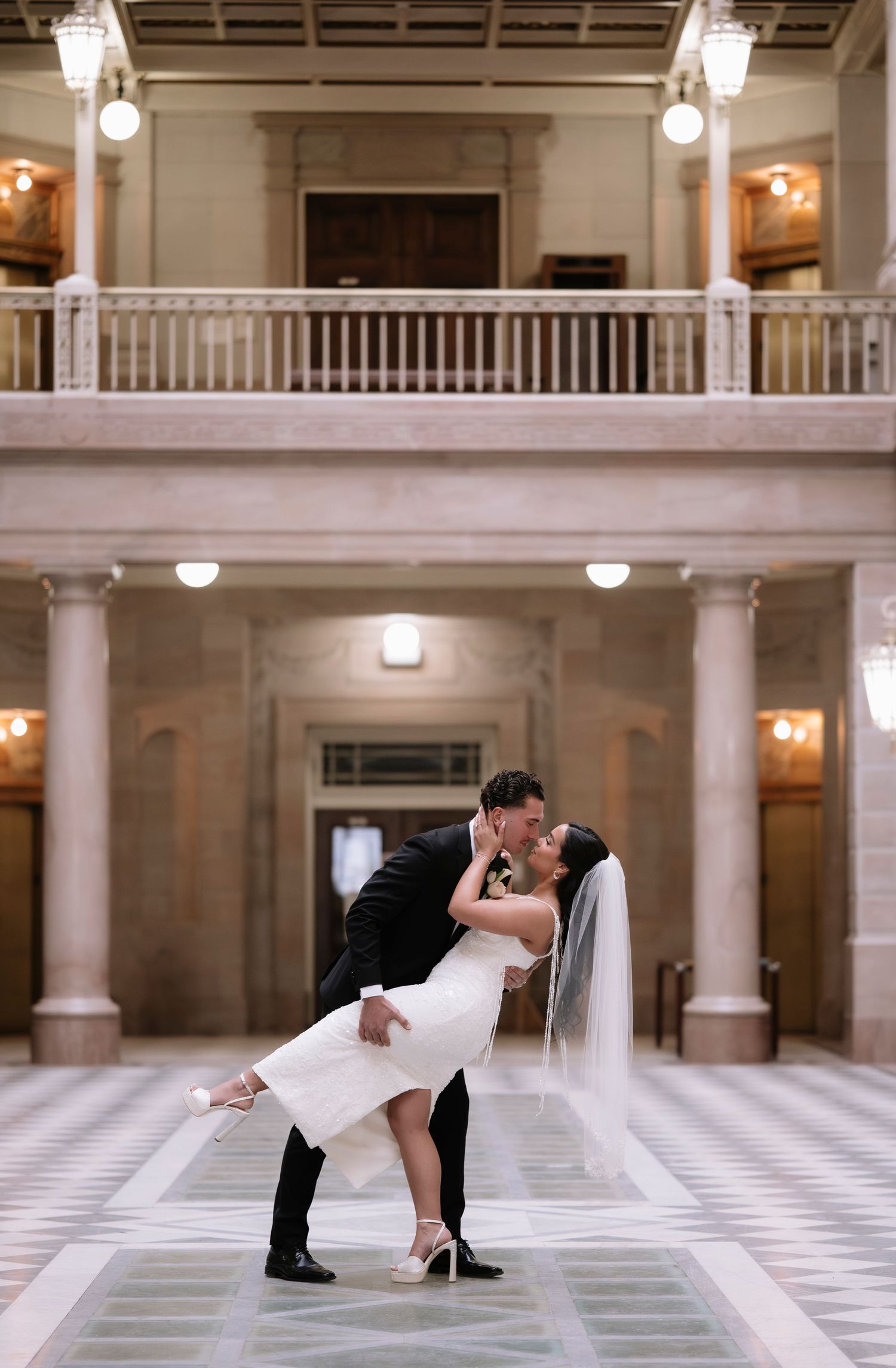 Couple sharing a romantic dip kiss in an elegant marble hall with columns and balcony during their wedding.
