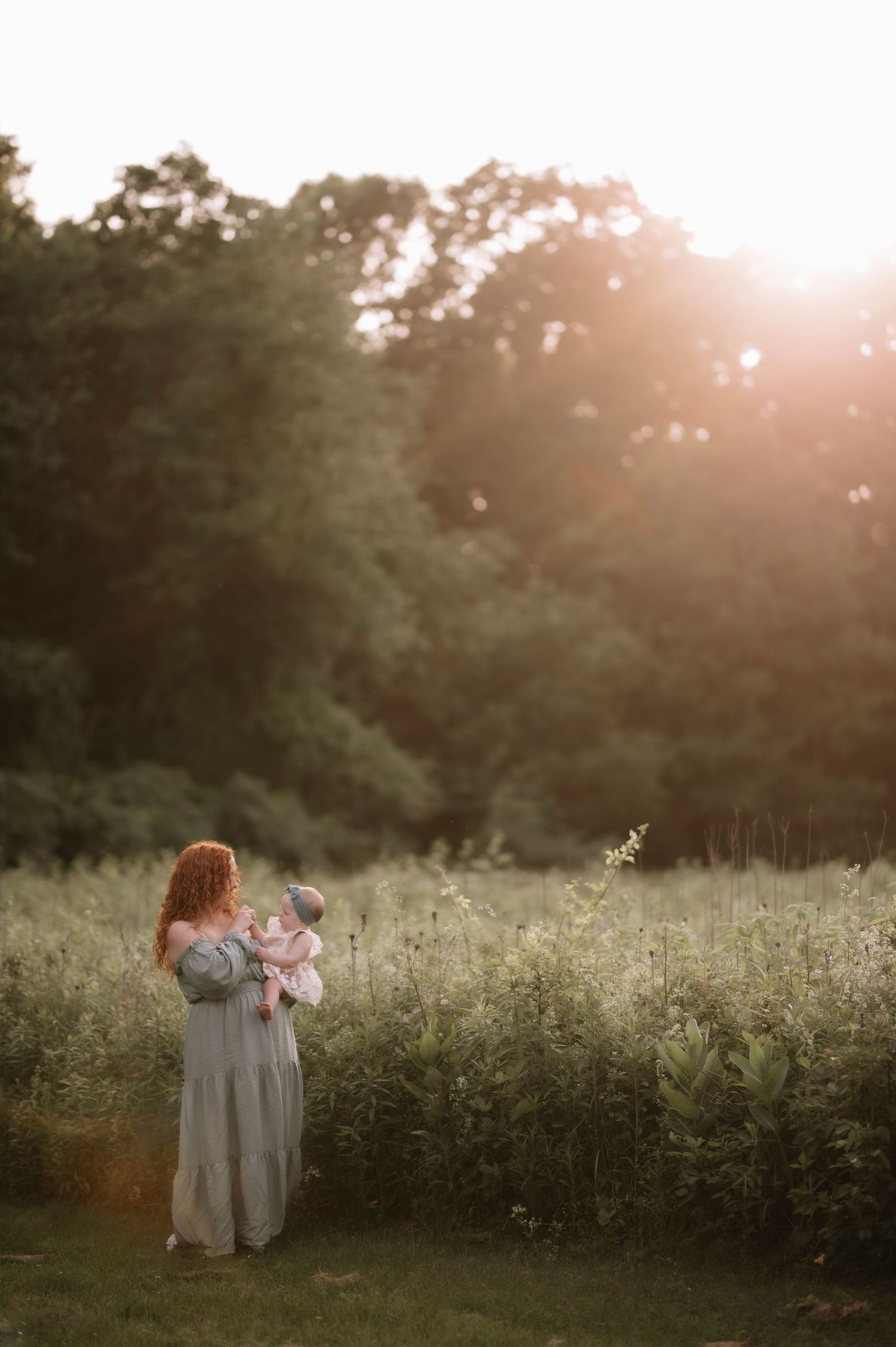 A woman in a flowy dress holds a baby in a misty meadow at sunset as golden light streams through the trees.
