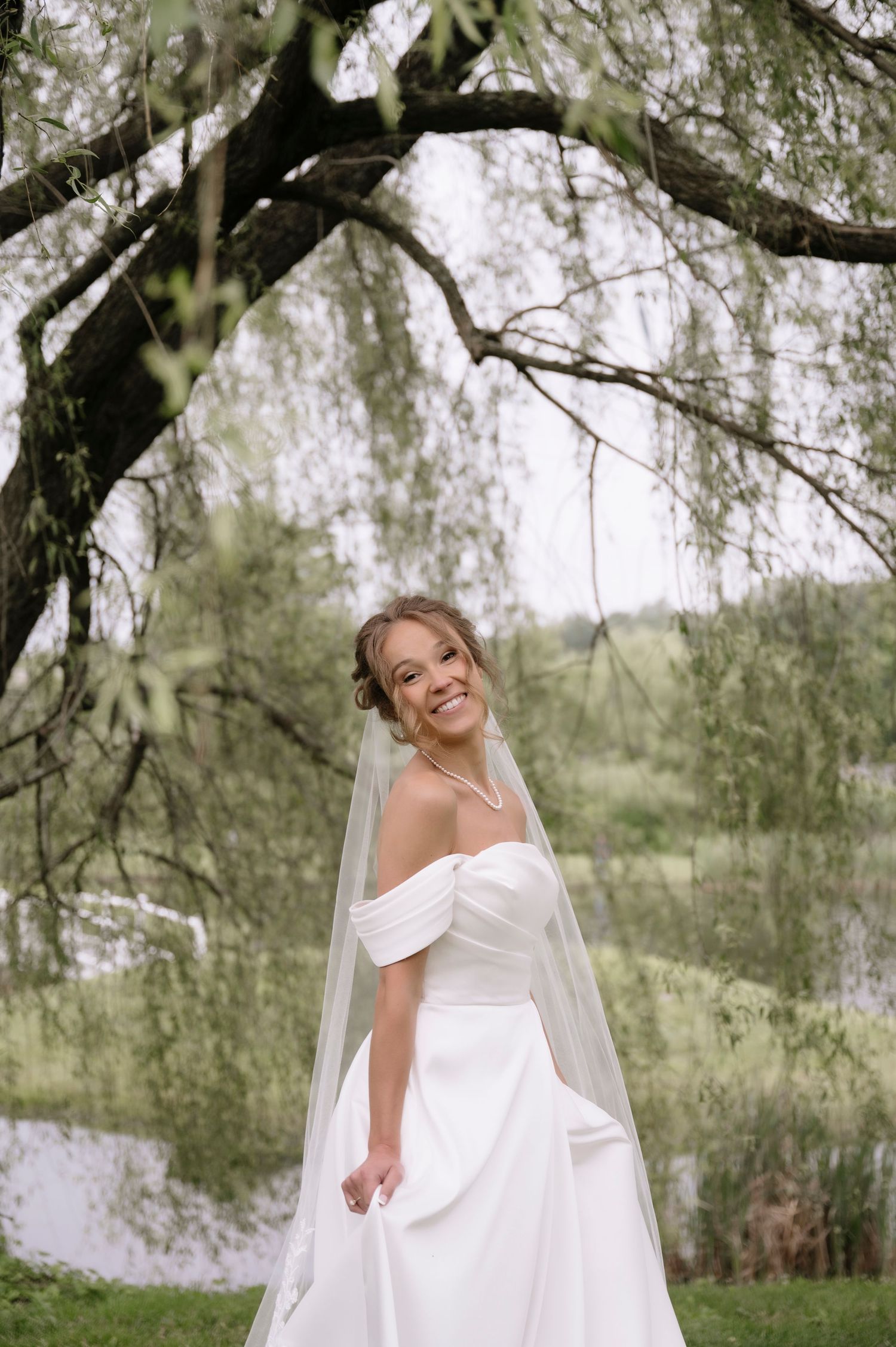 A bride in an elegant off-shoulder white wedding dress poses joyfully under willow trees near a lake.