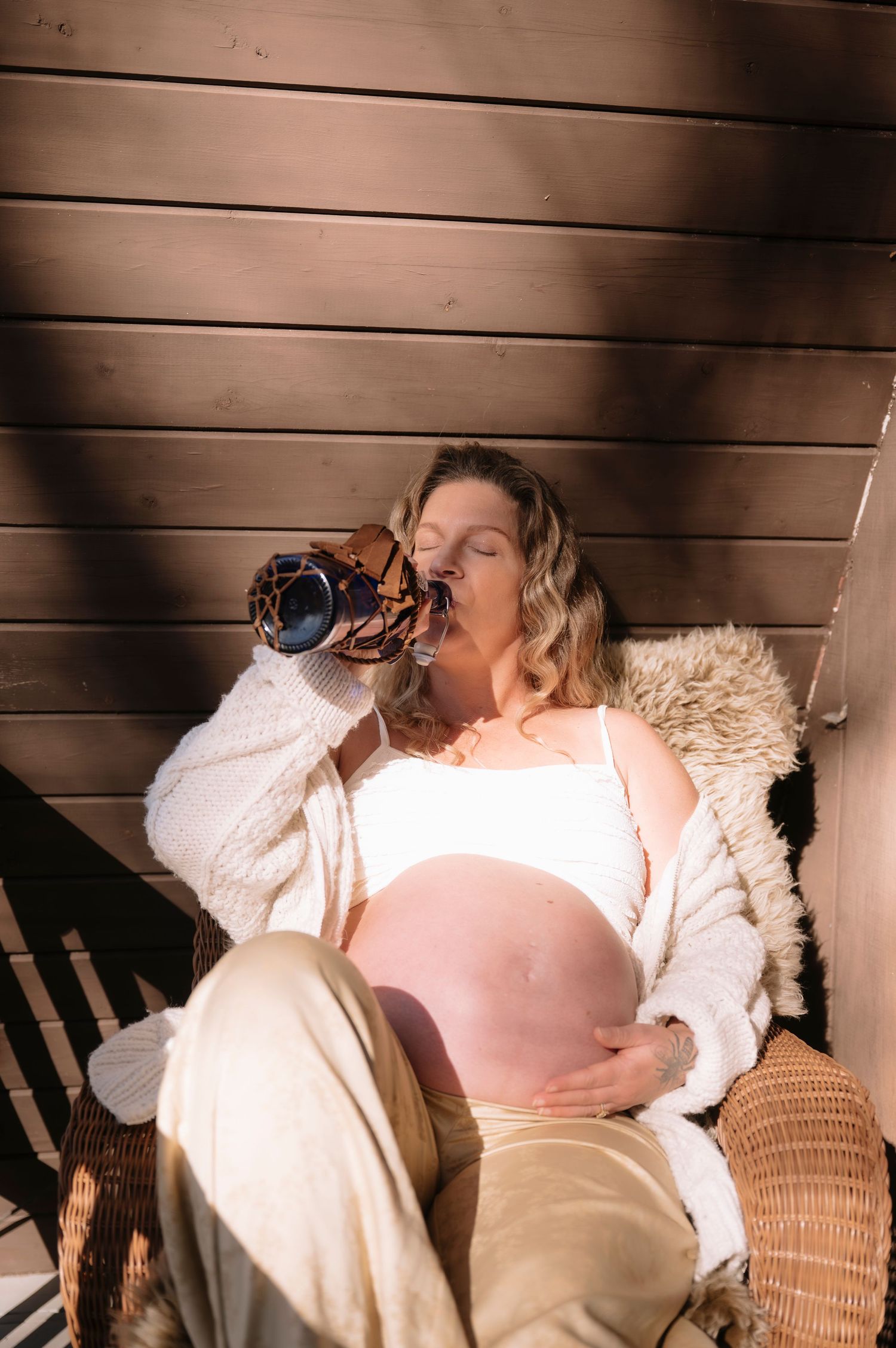 A pregnant woman in white loungewear drinks from a bottle while reclining on cushions against wooden walls.