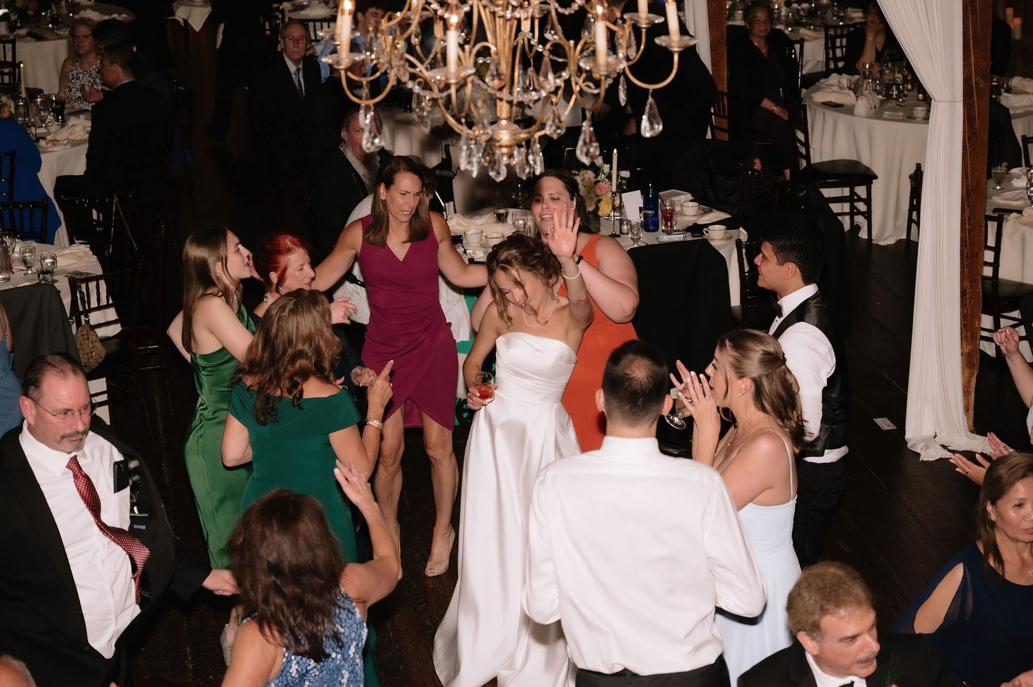 Bride in white gown dances joyfully with guests at elegant wedding reception under chandelier.