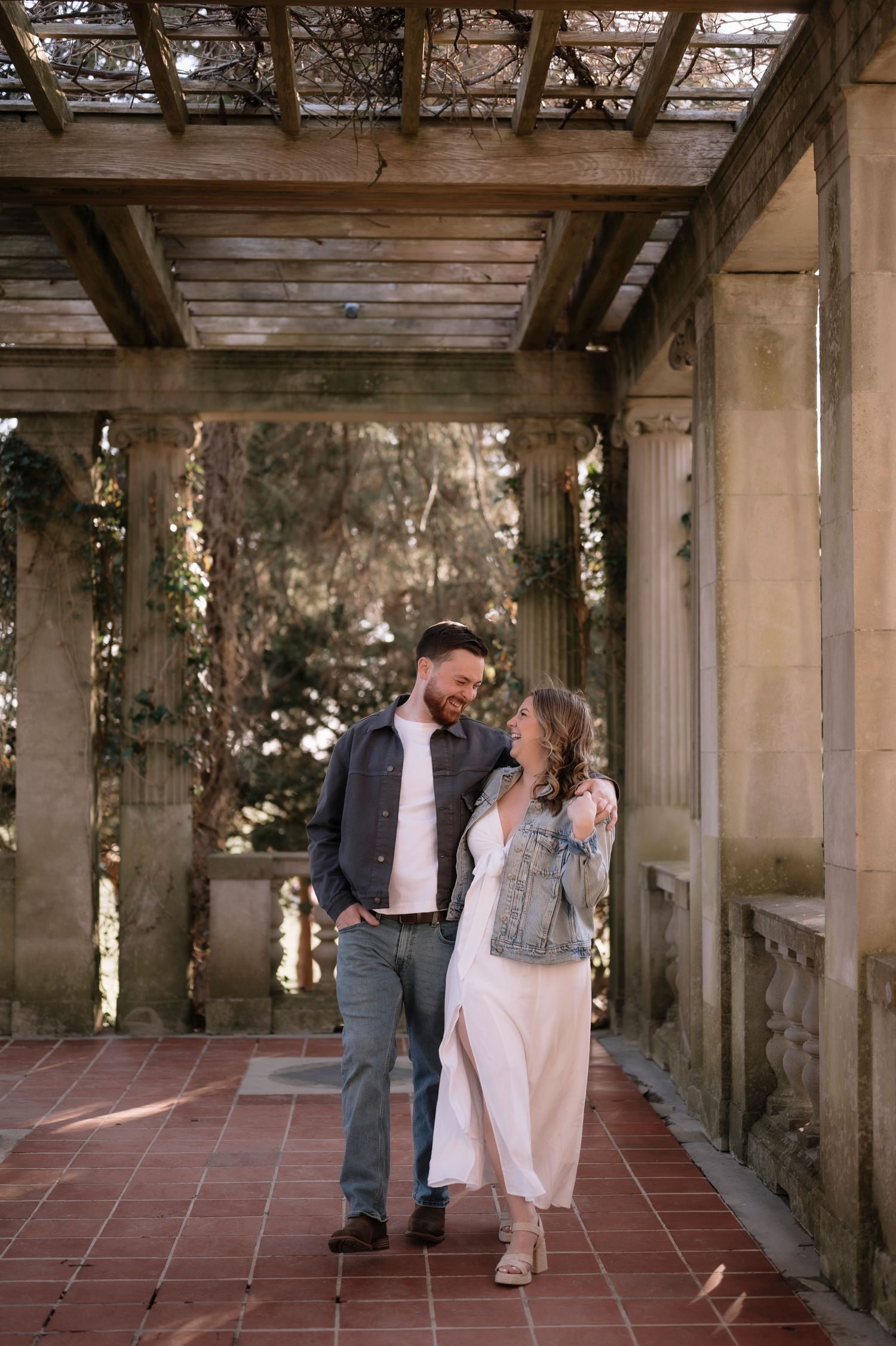 A couple shares a romantic moment under a rustic pergola with stone columns, she in a white dress and denim jacket, he in gray jeans.