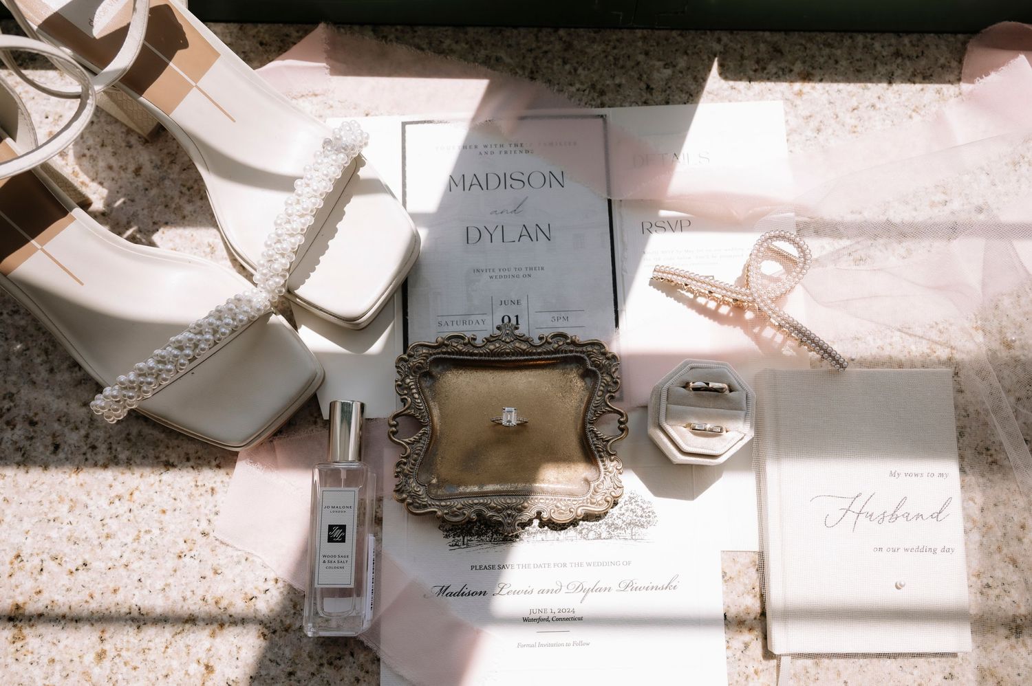Wedding details including invitation cards, pearl jewelry box, perfume bottle and ribbons arranged in soft sunlight.