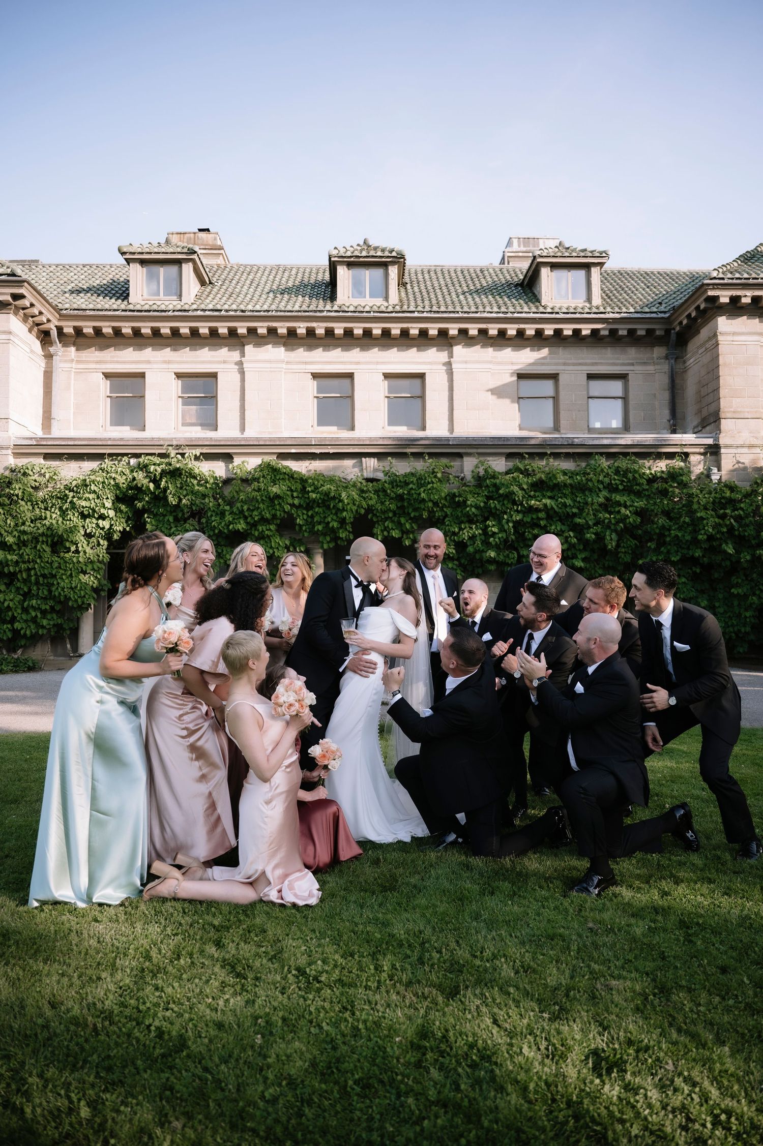Wedding party poses together in a playful group photo on lawn in front of elegant stone mansion.