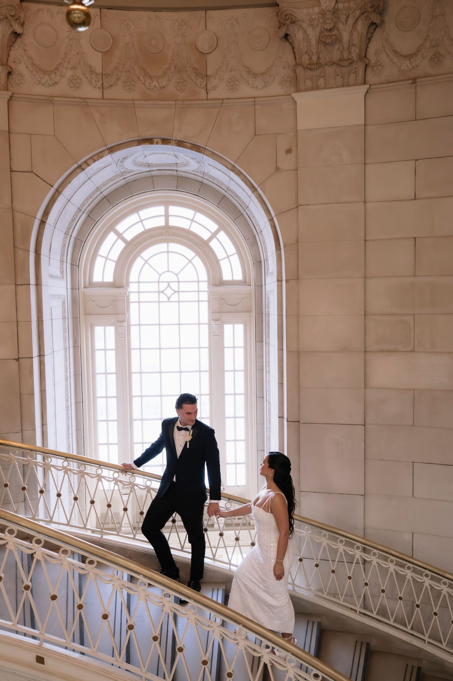 Bride and groom hold hands on elegant staircase with arched window at San Francisco City Hall wedding.