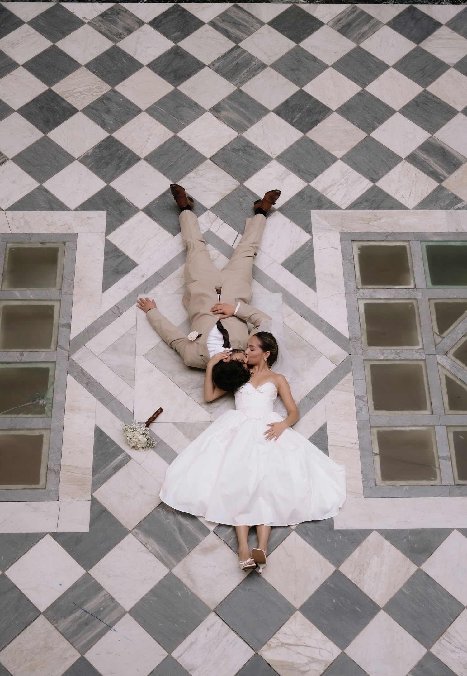 A couple in wedding attire lies on a geometric black and white checkered floor with glass panels.