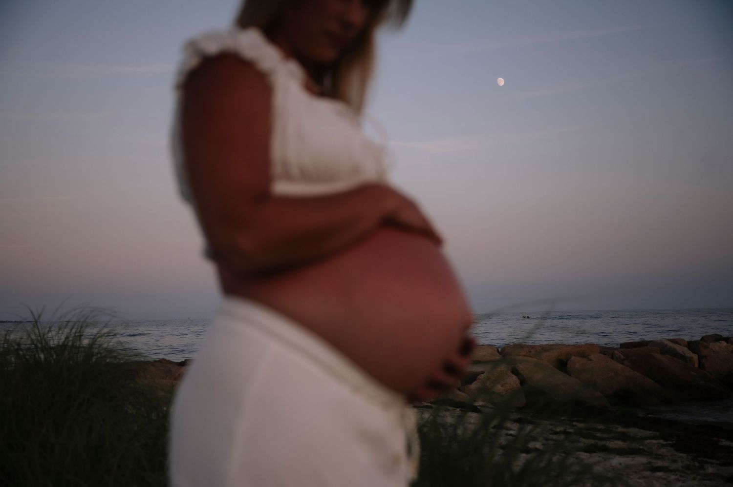 Pregnant woman in white dress cradling baby bump at beach during twilight with moon visible in sky.